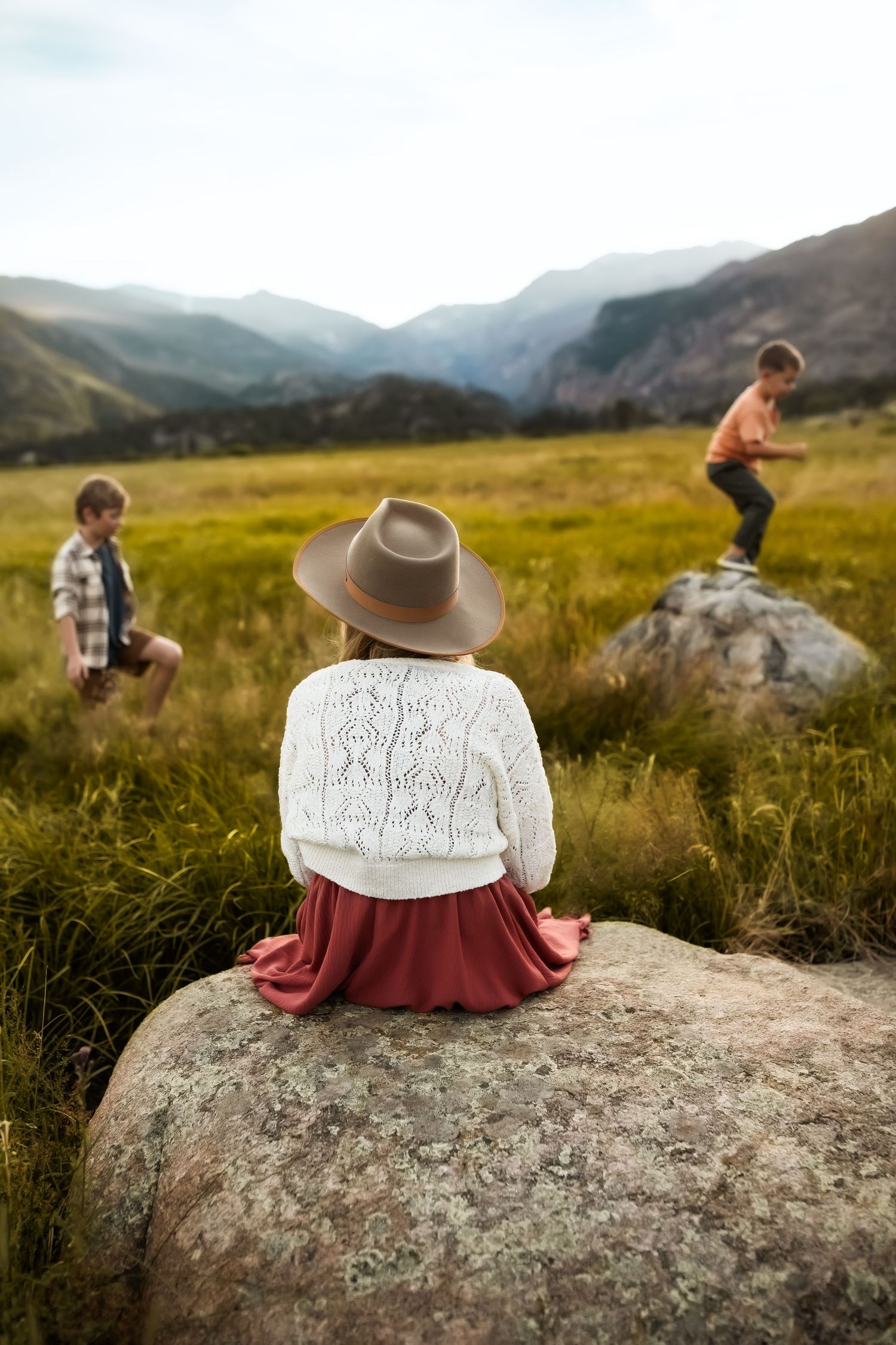 A person wearing a hat sits on a large boulder overlooking scenic mountain meadows at sunset.