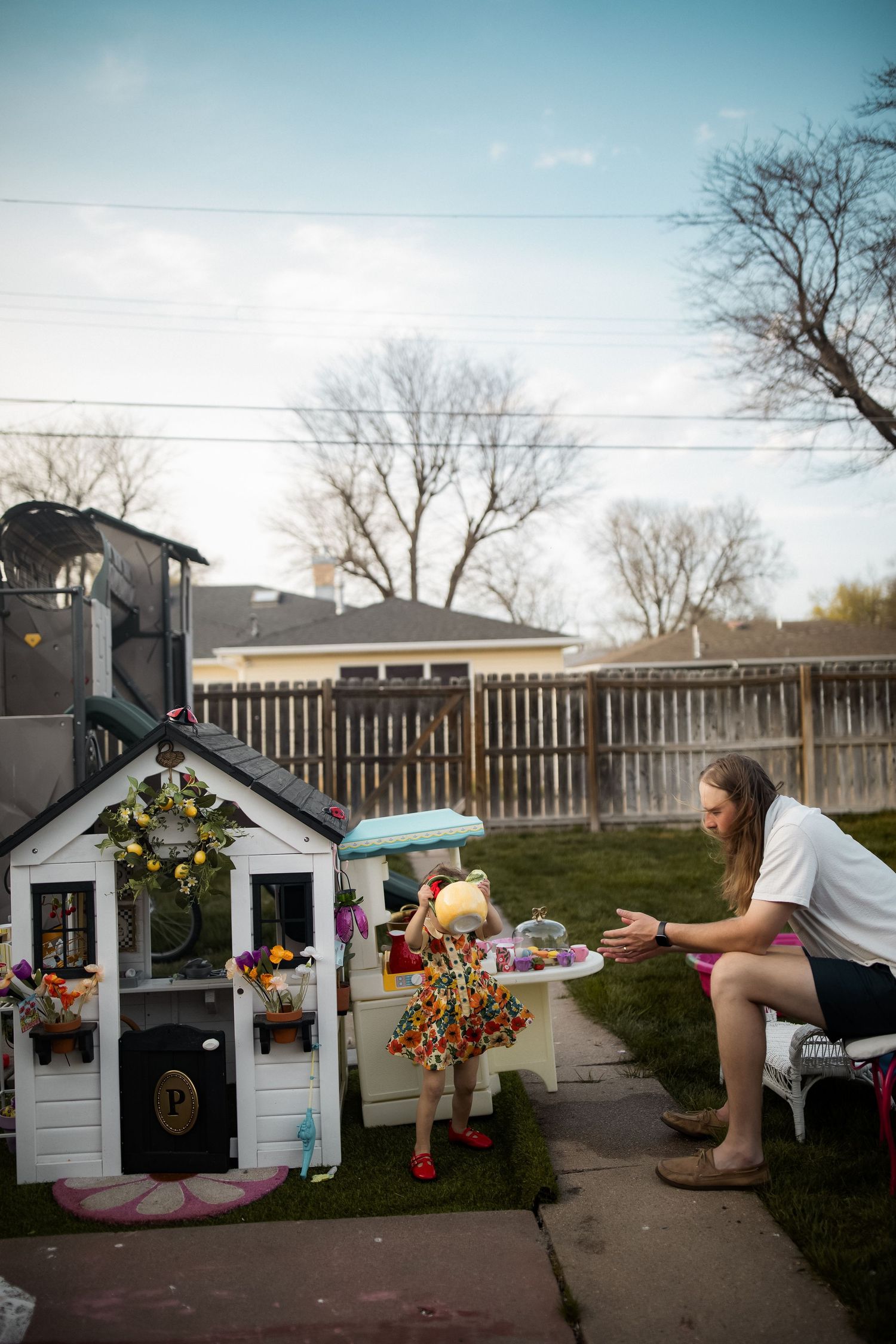 A man crouches to interact with a young girl playing pretend outside a toy playhouse in a backyard.