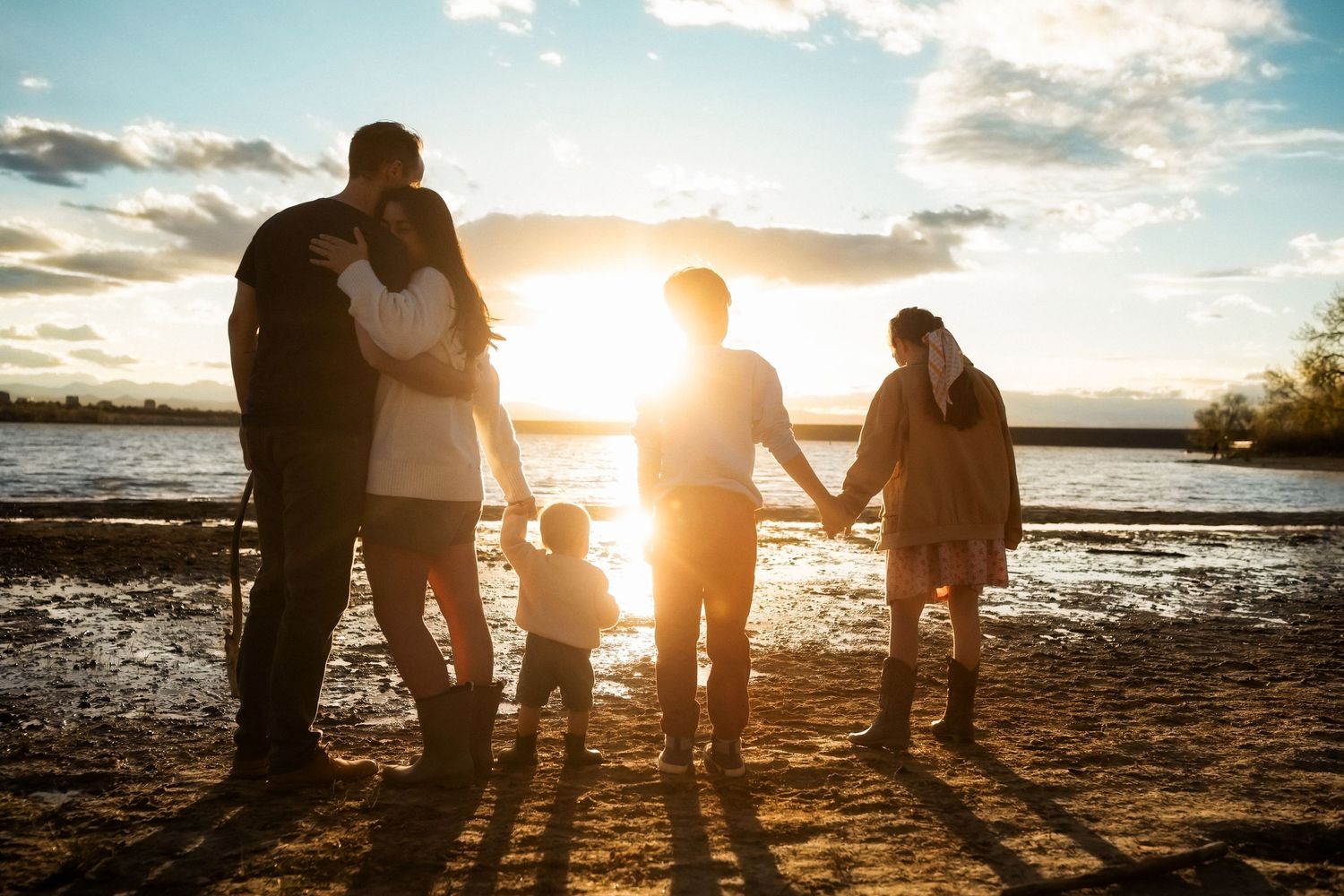 Family silhouetted against a golden sunset on a sandy beach shore, holding hands together.
