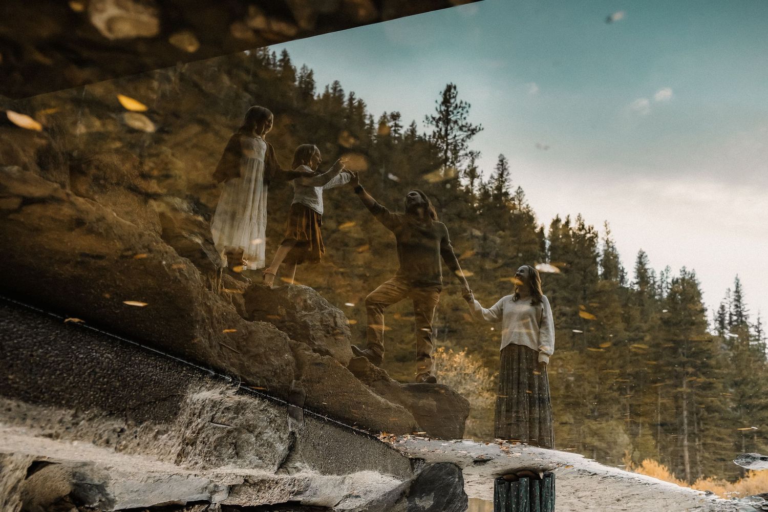 Reflection of people standing on rocks near pine trees captured in a puddle with golden leaves in a mountain setting.