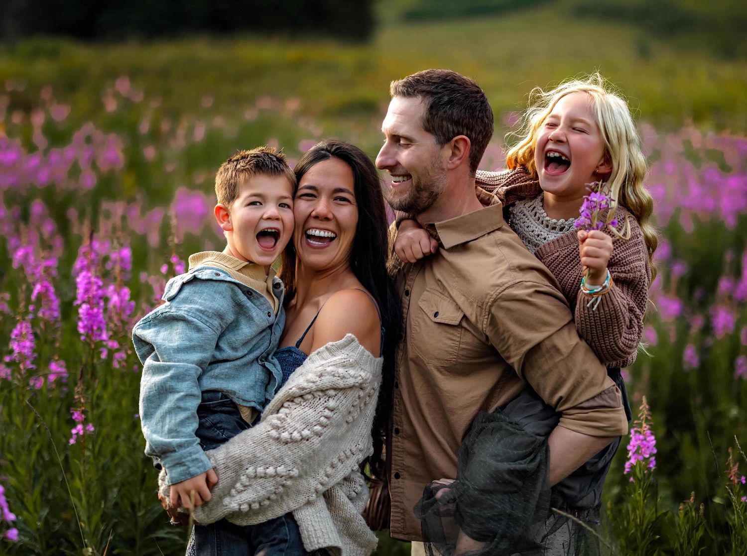 Happy family of four laughing in a vibrant wildflower meadow, parents holding children during outdoor photo session.