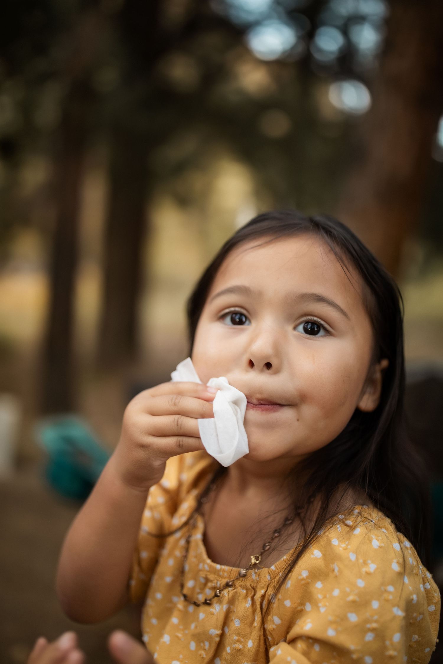 Child in polka dot dress eating a marshmallow outdoors with trees in background.