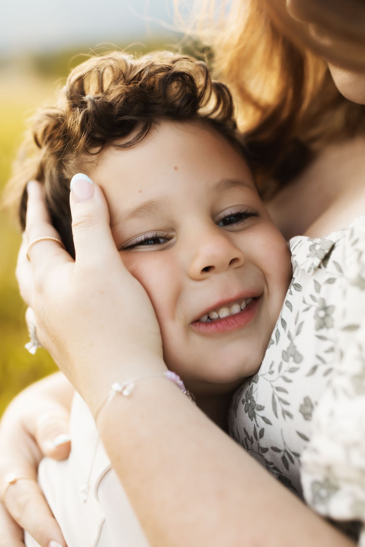 A child with curly hair smiles warmly while being embraced in a tender moment outdoors.