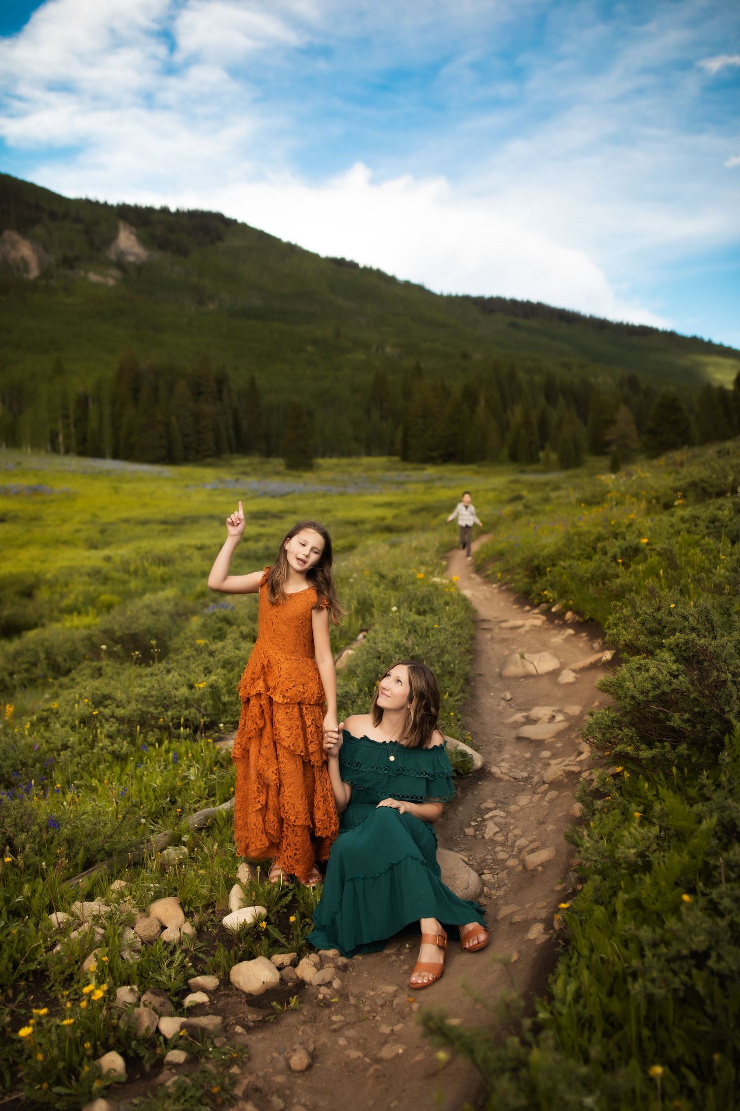Two people in colorful dresses sitting on a mountain trail surrounded by wildflowers.