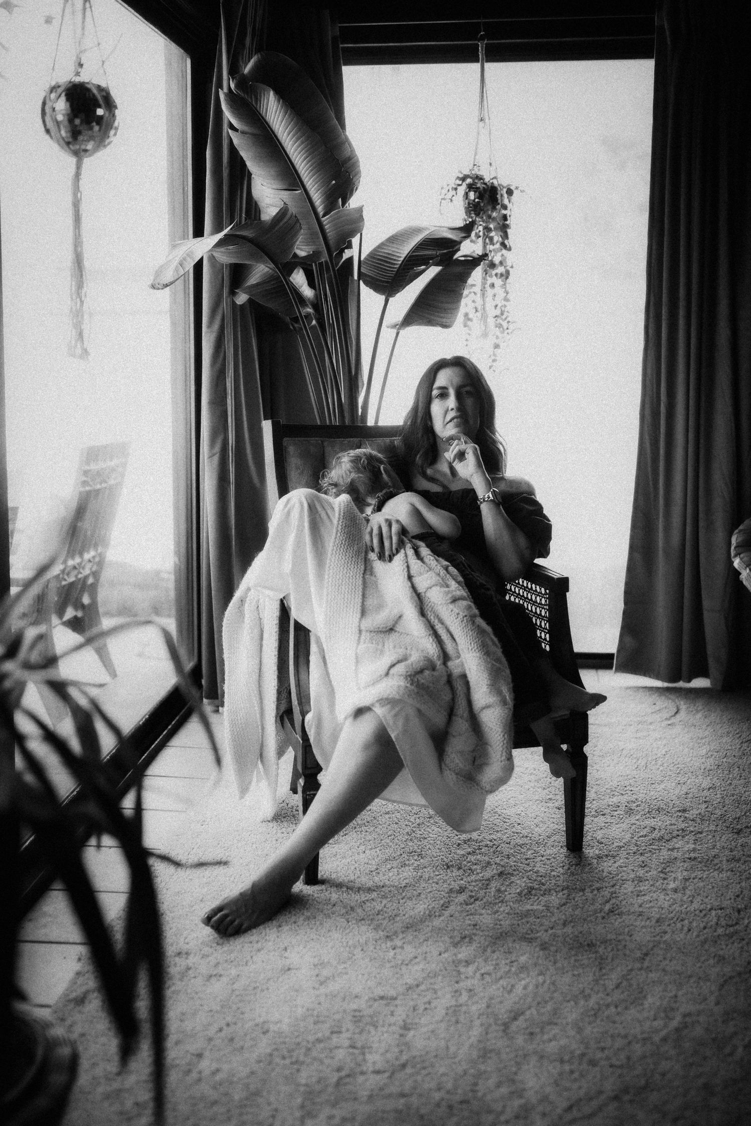 Black and white photo of a woman sitting in a chair wrapped in a fur blanket surrounded by indoor plants.