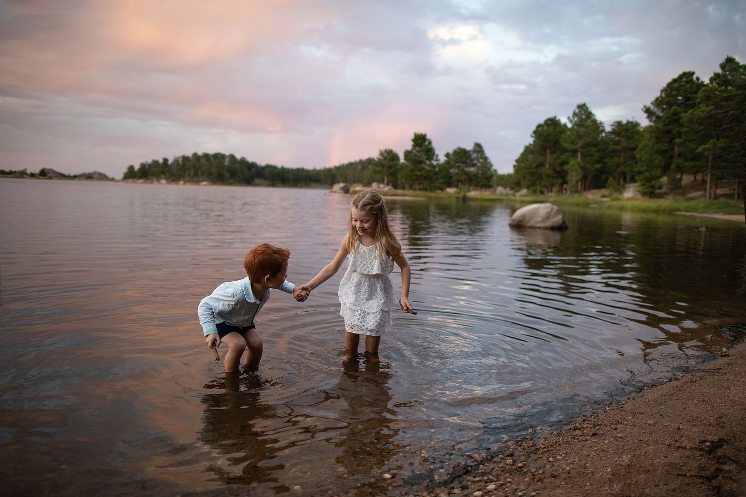 Sunset lakeside scene with people exploring shallow waters near pine-covered shore.