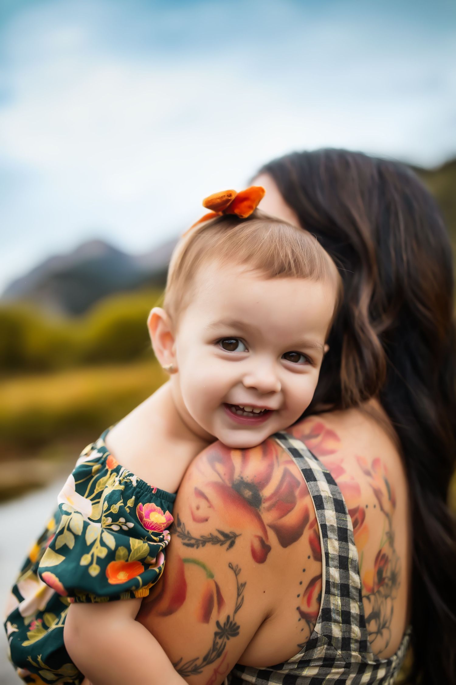 Baby wearing floral print outfit smiling while being held near colorful autumn scenery.