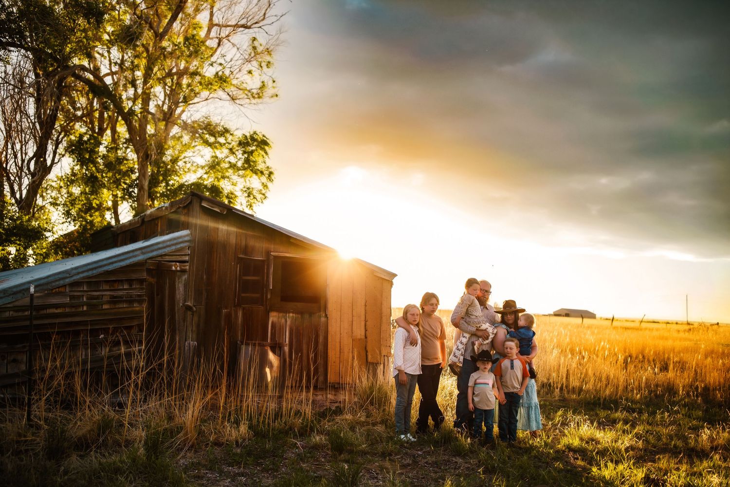 Silhouettes of people stand near a rustic barn at sunset in a golden wheat field with dramatic sky.