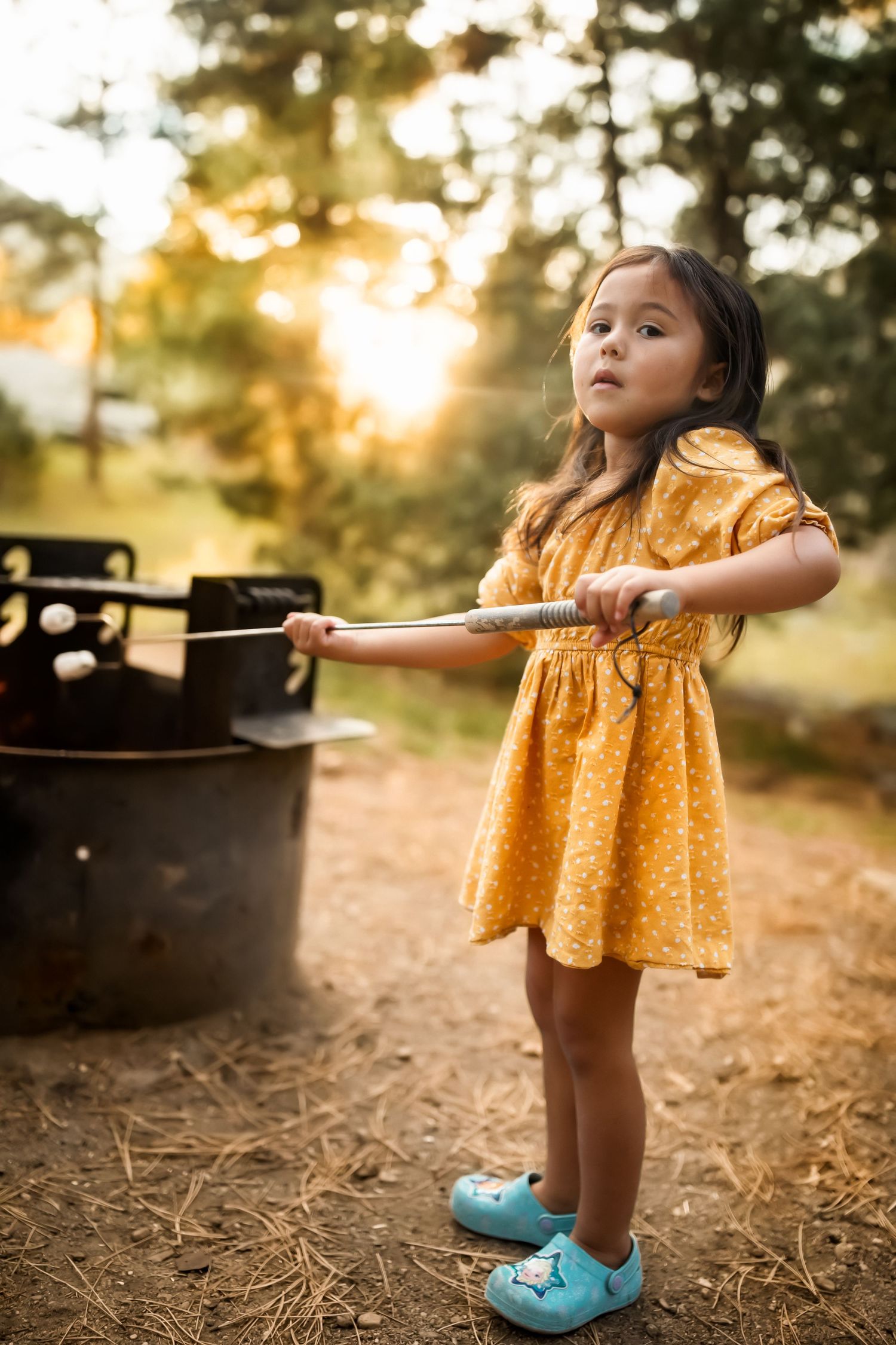 A small child in a yellow dress and blue shoes stands in dappled sunlight on a forest floor.