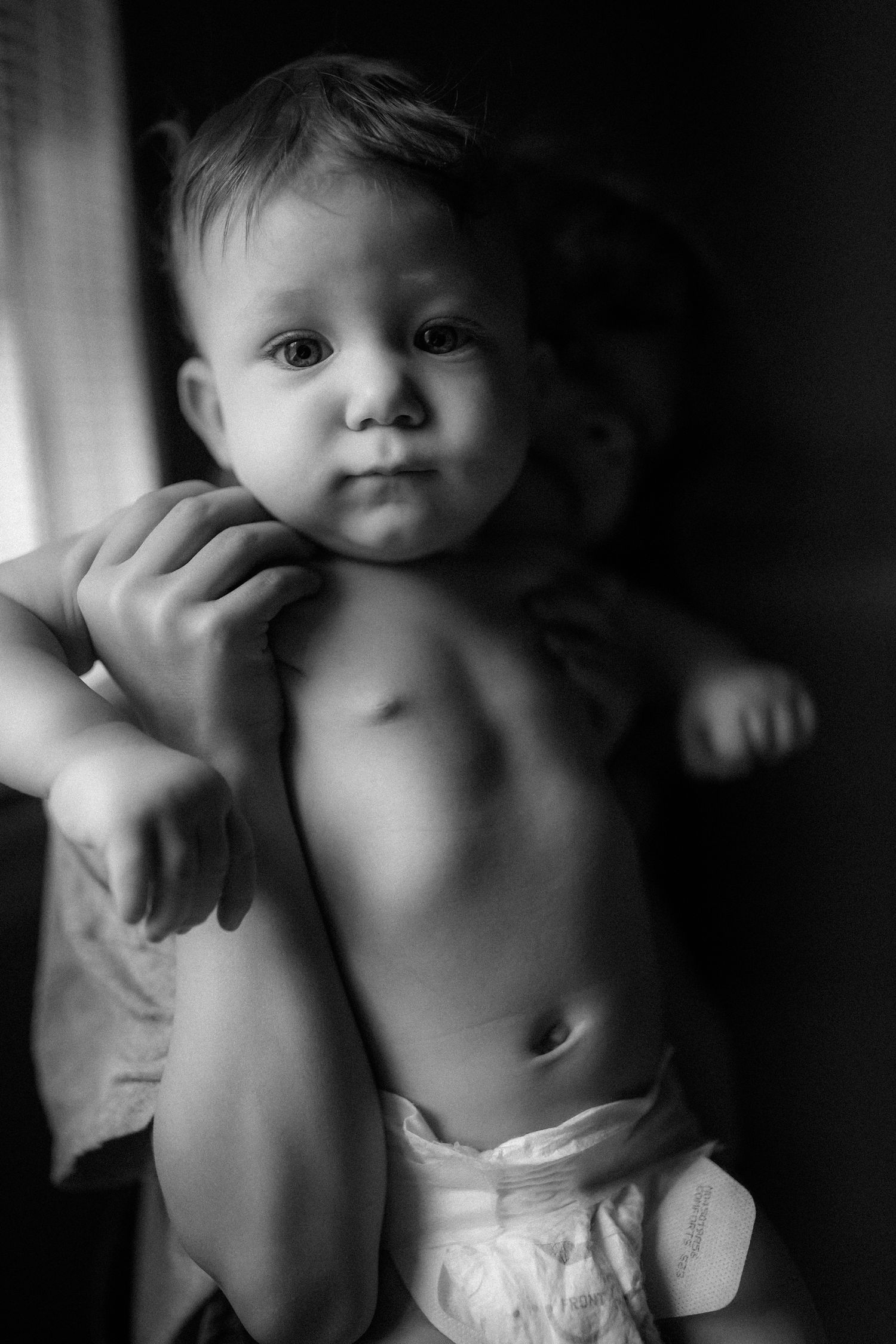 A black and white portrait of a toddler in a diaper near a window.