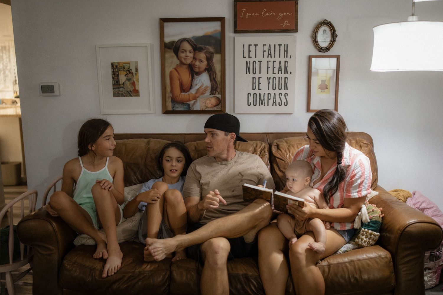 Family relaxes together on brown leather couch in cozy living room with framed artwork on walls.