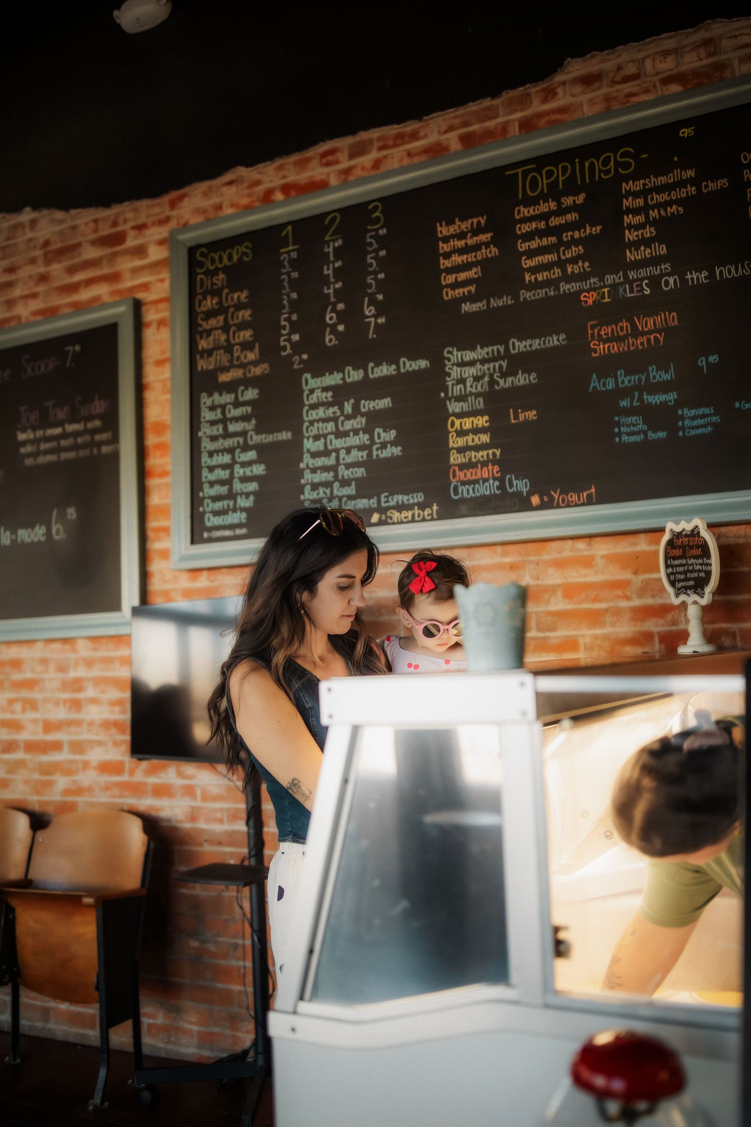Woman ordering at an ice cream shop counter with a chalkboard menu on a brick wall.