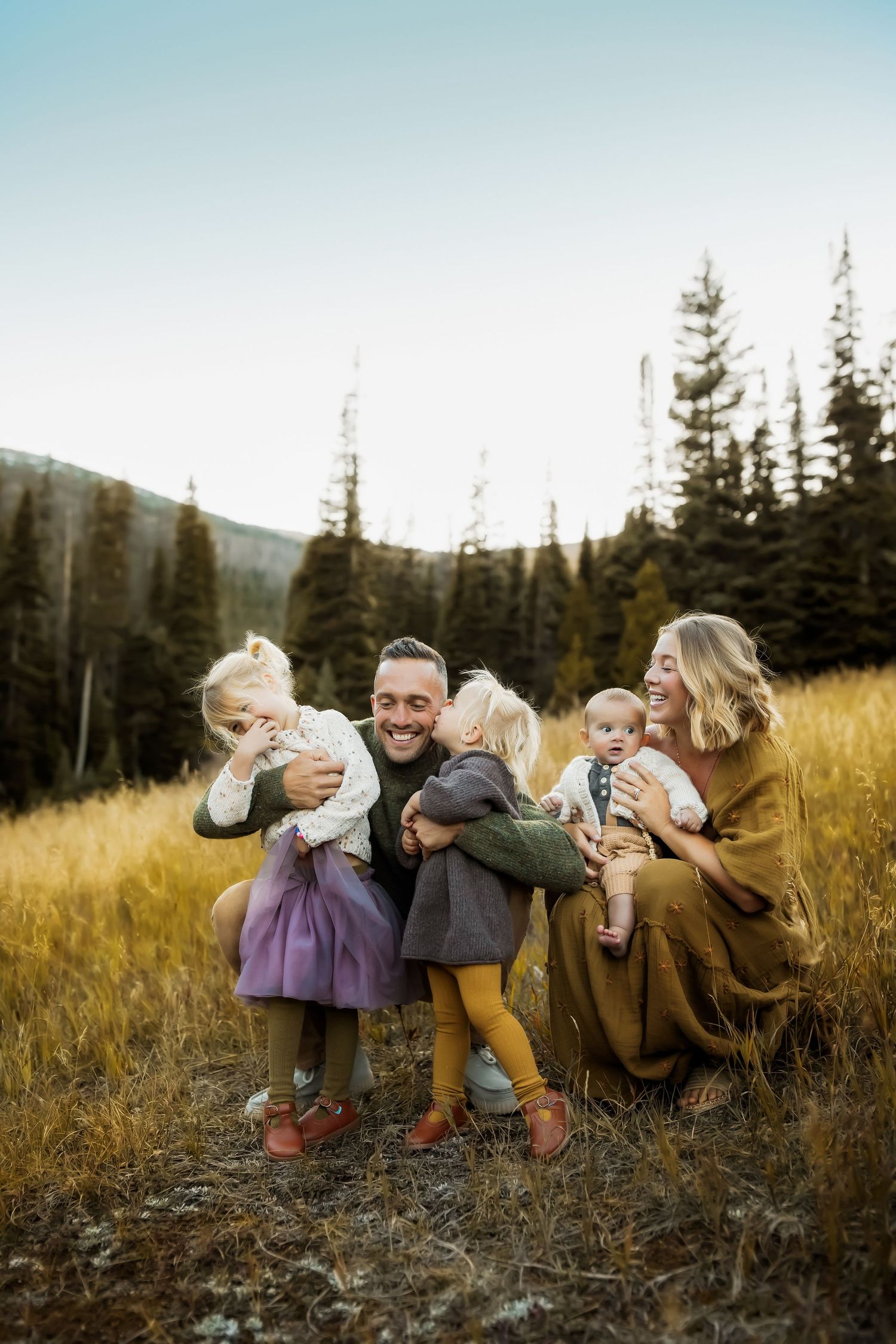 A playful group sits in a golden field at sunset surrounded by evergreen trees in the mountains.