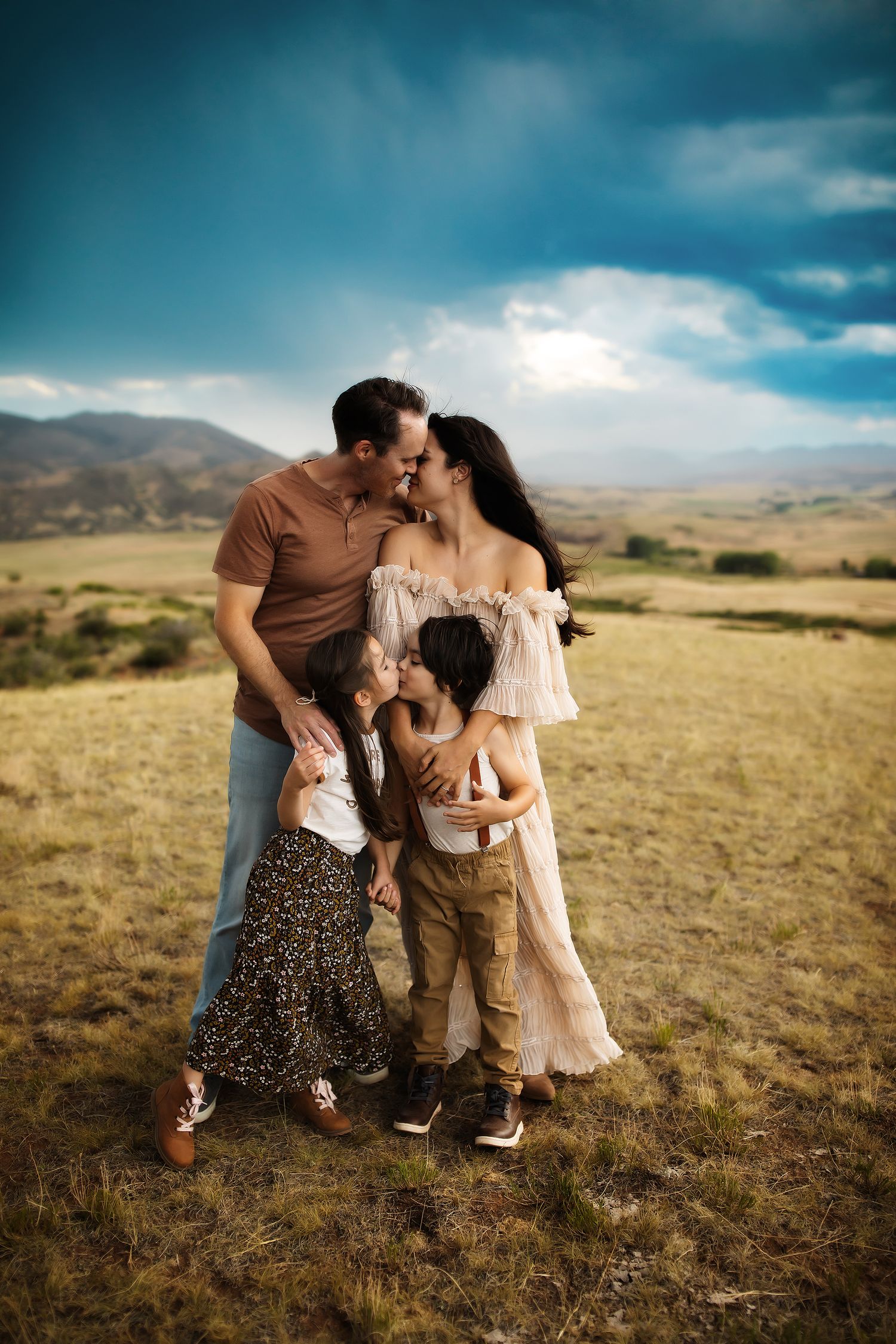 Family embracing together on a golden hillside with dramatic cloudy skies and mountains in the background.