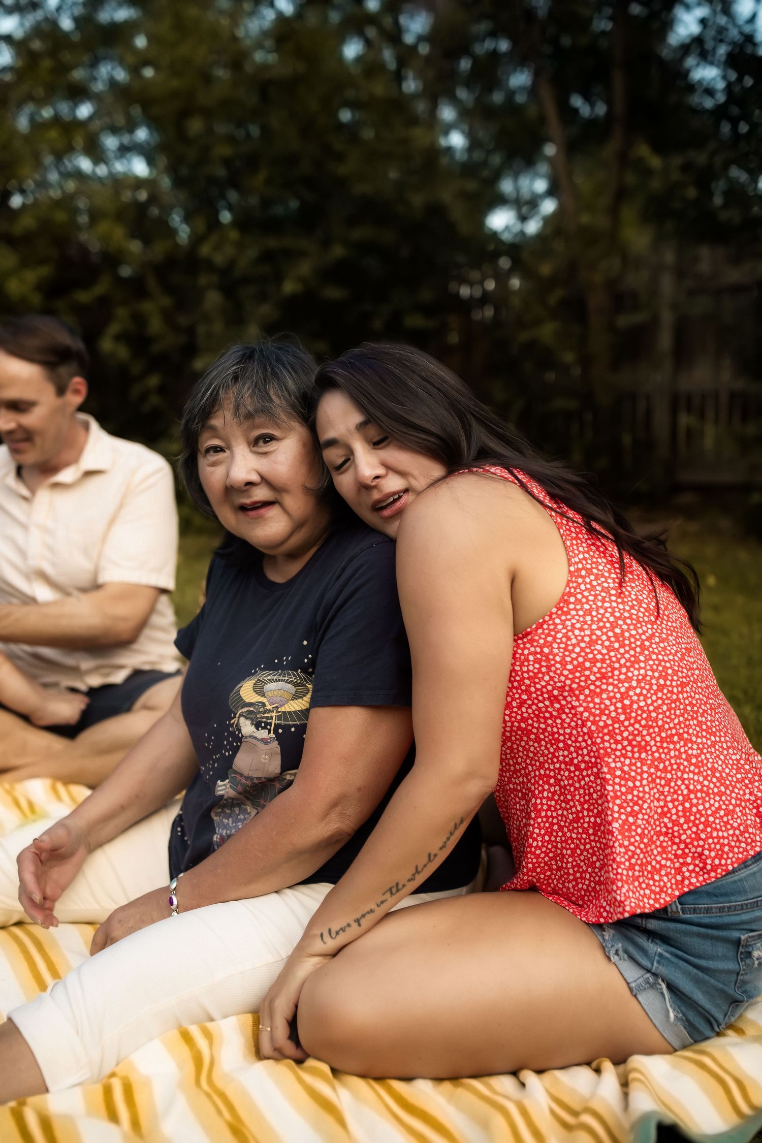 A couple shares a tender moment while lying on a striped blanket outdoors in a natural setting.