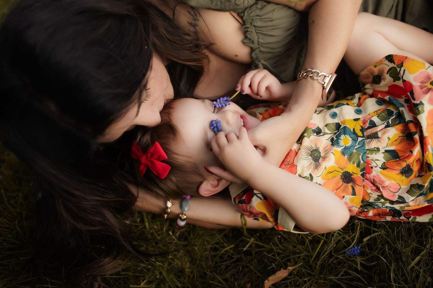 A mother tenderly kisses her young daughter lying on green grass, both wearing colorful floral outfits.