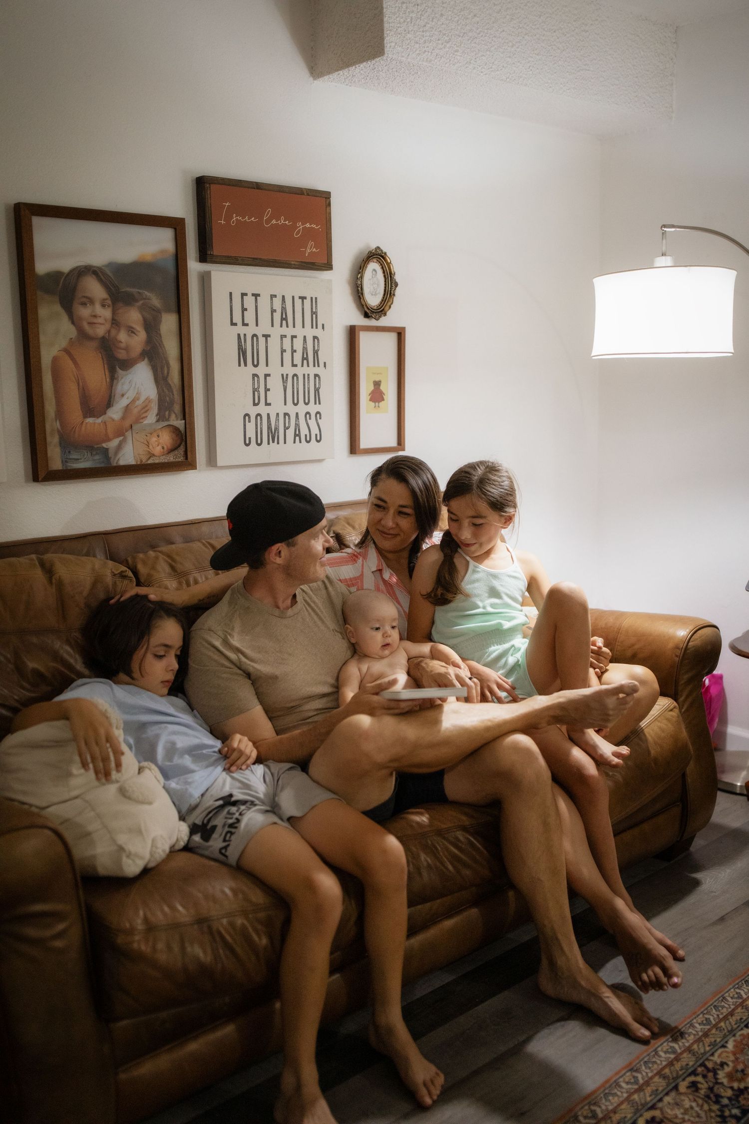 A family relaxes together on a brown leather couch in a cozy living room with framed wall art.
