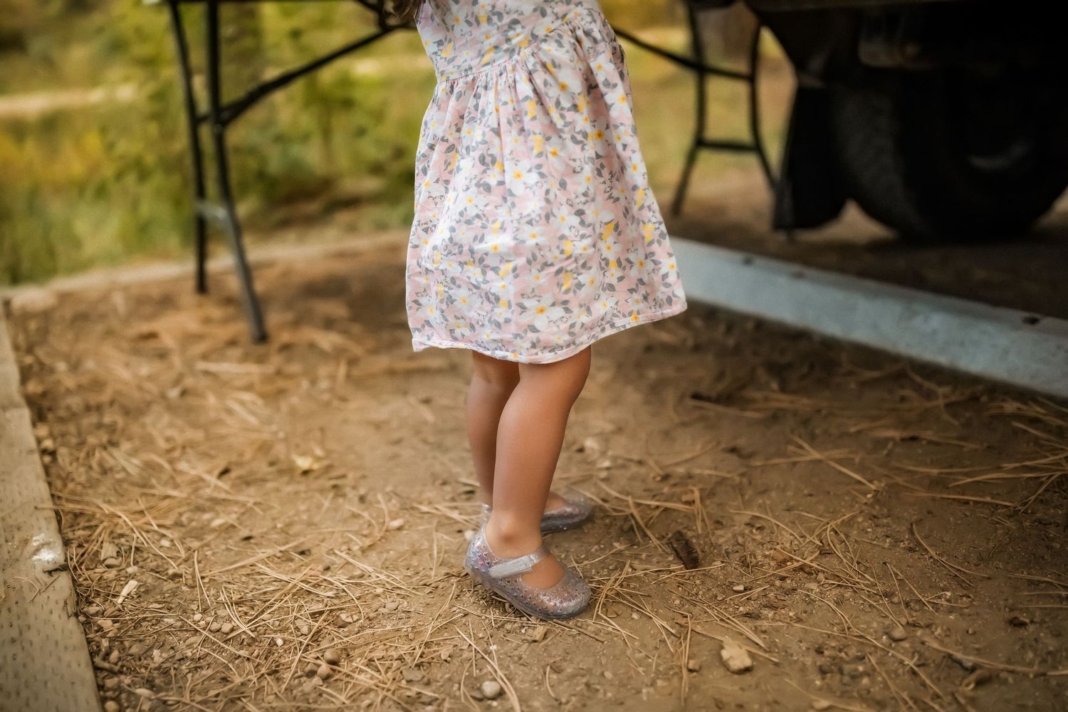 Small feet in gray shoes stand on dirt ground wearing a floral patterned dress on a sunny day.