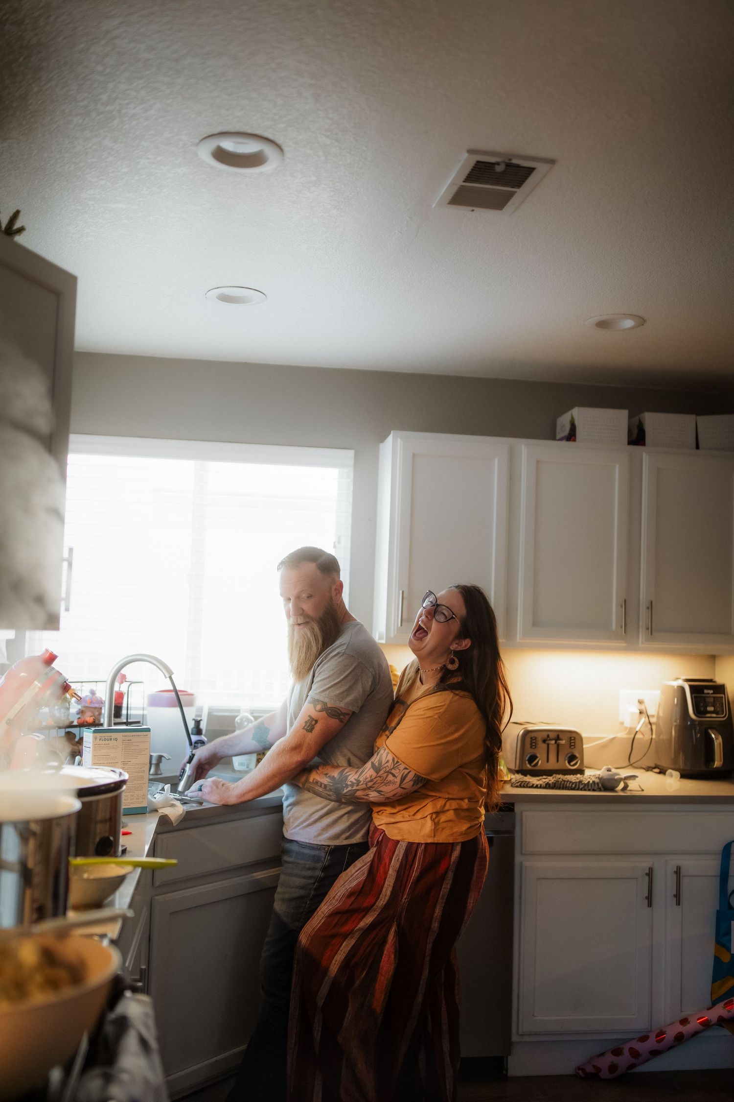 A couple shares a tender moment in a bright kitchen, with the man washing dishes and the woman embracing him from behind.