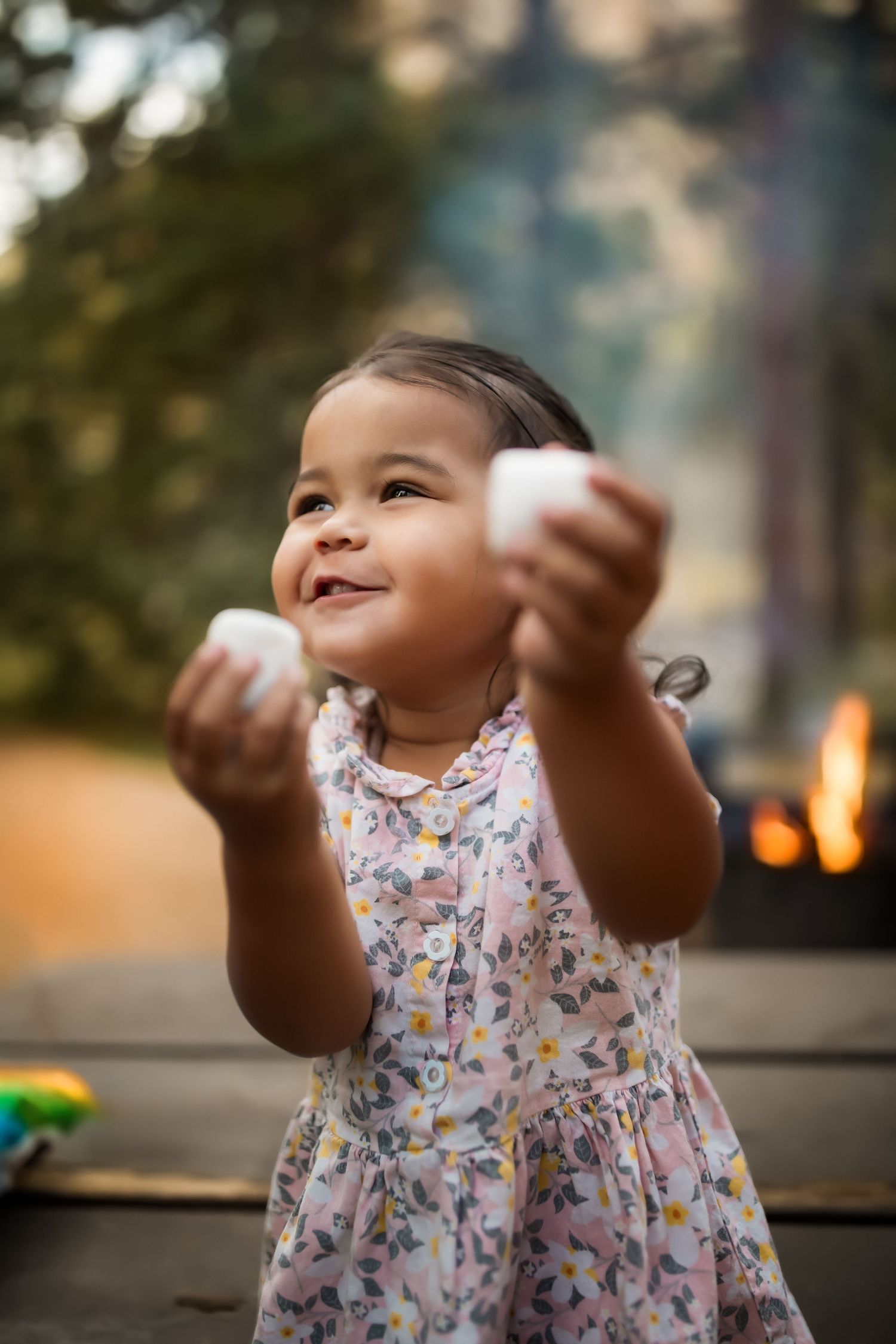 Young child in floral dress joyfully playing with marshmallows outdoors at dusk.