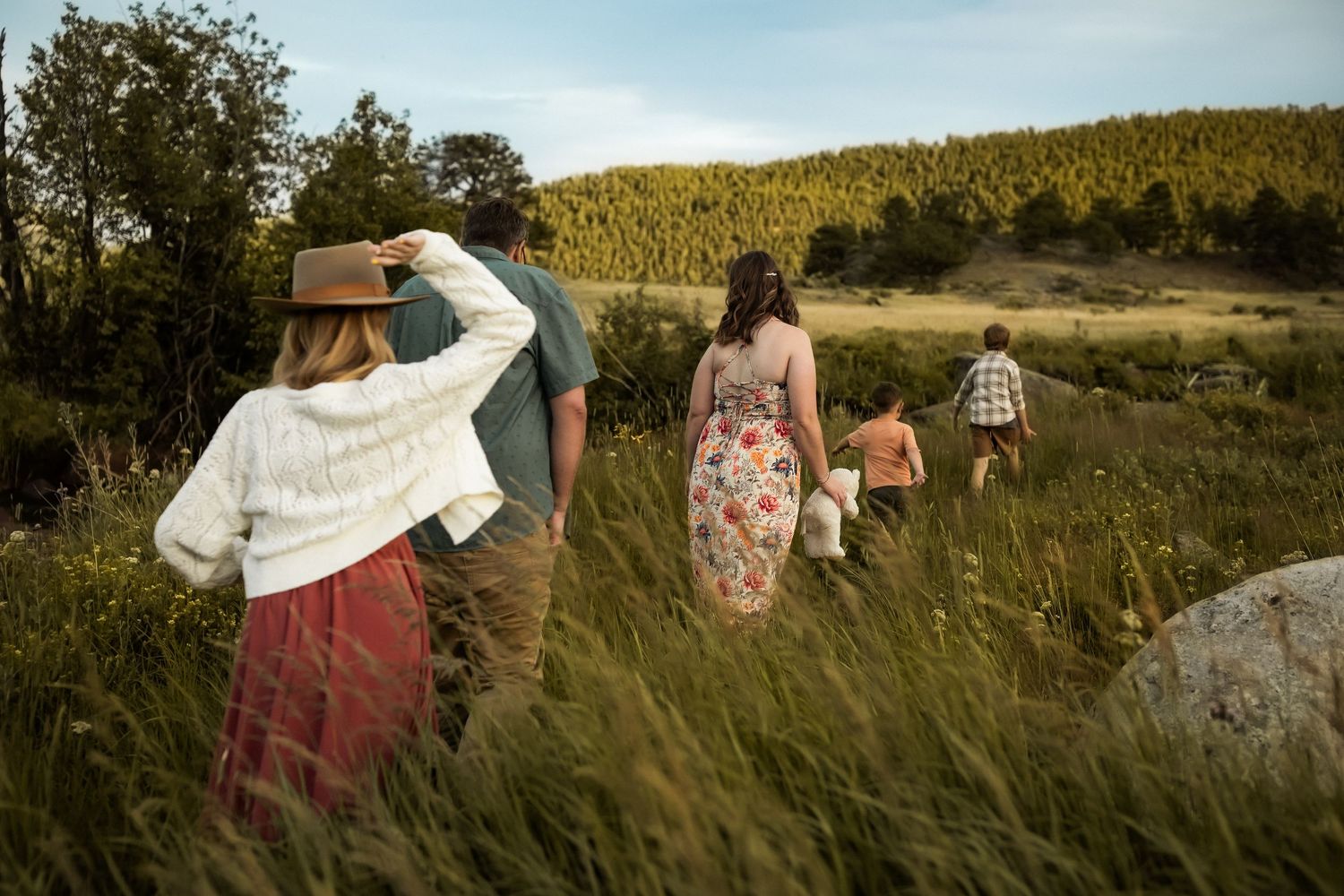 A group walks through a tall grass meadow at golden hour with mountains and trees in the background.