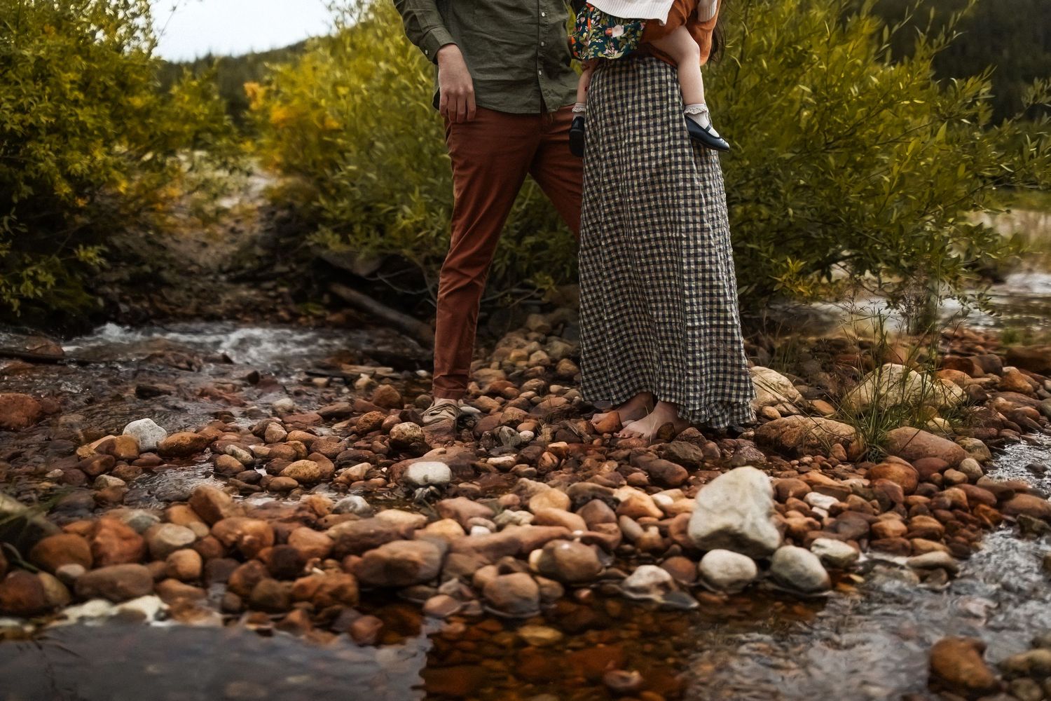 People exploring rocky riverbank with mountains and fall colors in background.