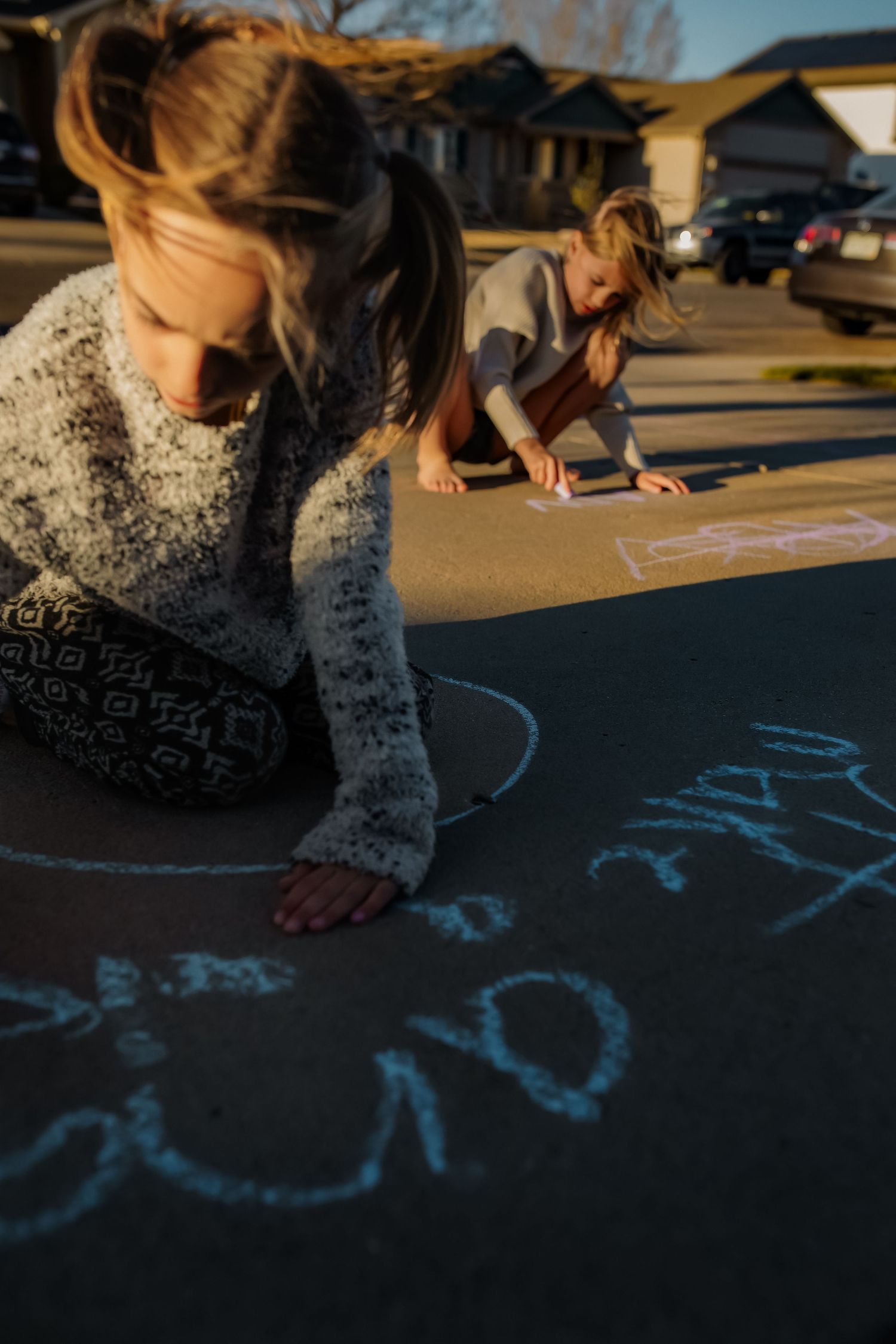 Two children drawing colorful chalk art on a suburban driveway in warm evening sunlight.