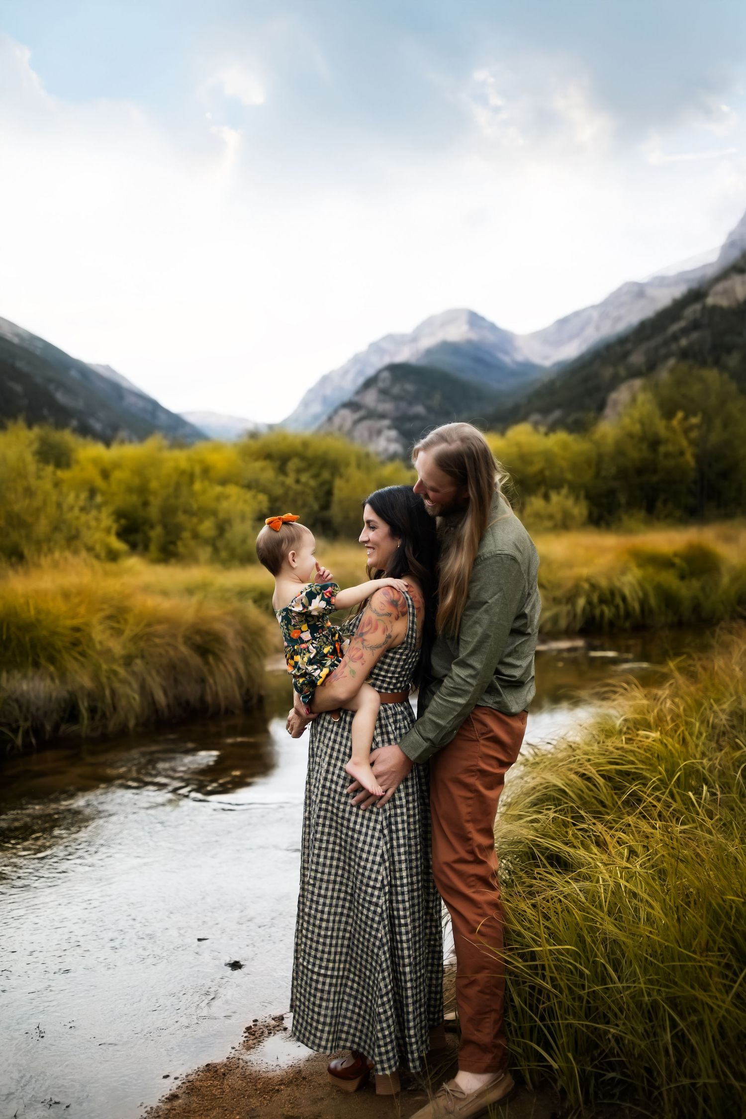 A family stands together by a mountain stream with dramatic peaks and autumn foliage in the background.