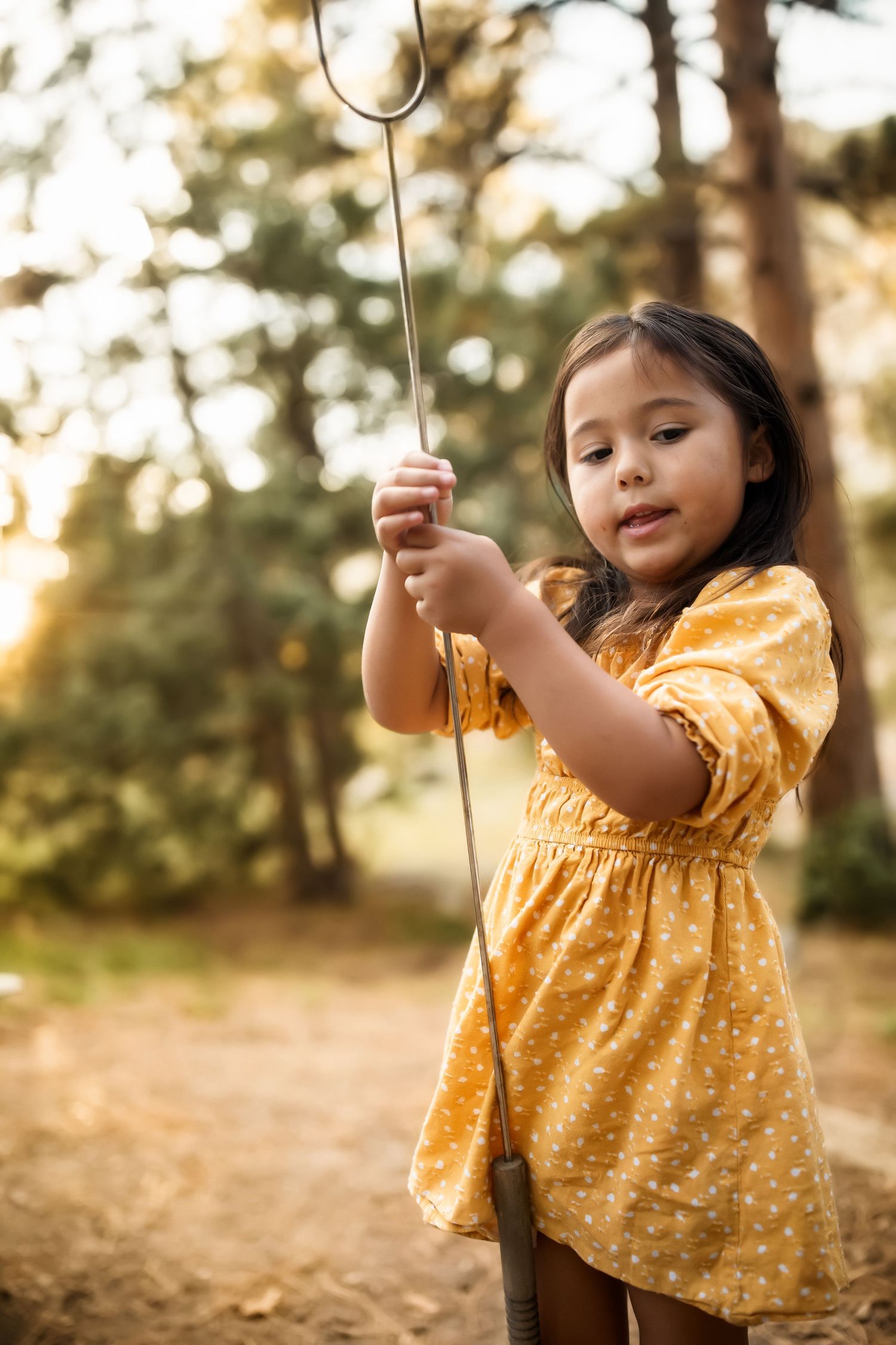 Child in yellow polka dot dress holding roasting sticks outdoors.