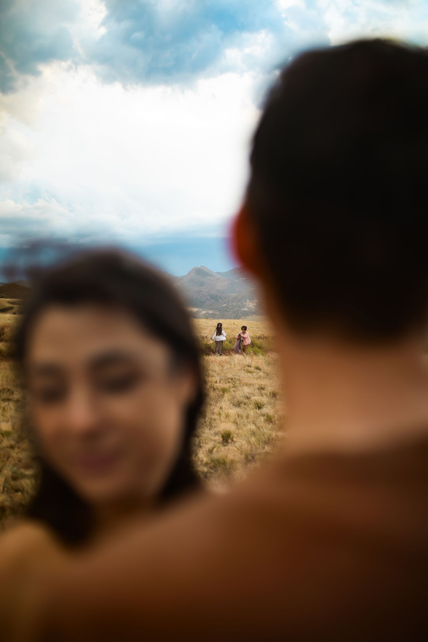Couple in foreground with two people walking in golden field under dramatic cloudy sky and mountain backdrop.