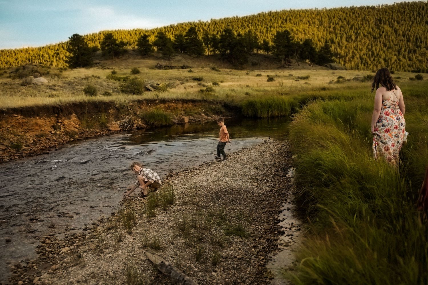 People walking along a rocky riverbank trail during sunset in a mountainous landscape.
