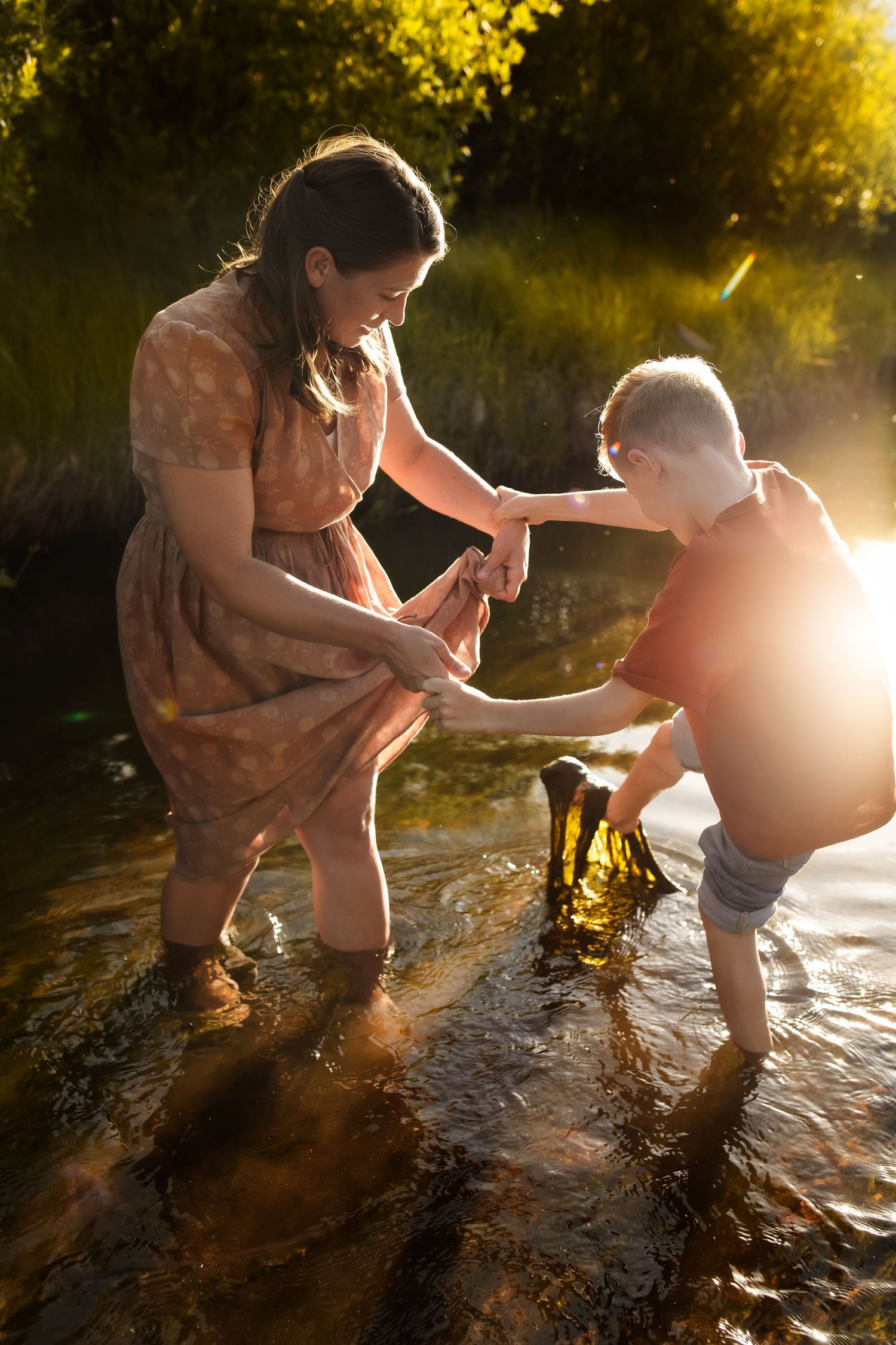 Woman in floral dress helps boy wade through sunlit river, golden hour light glowing through trees.