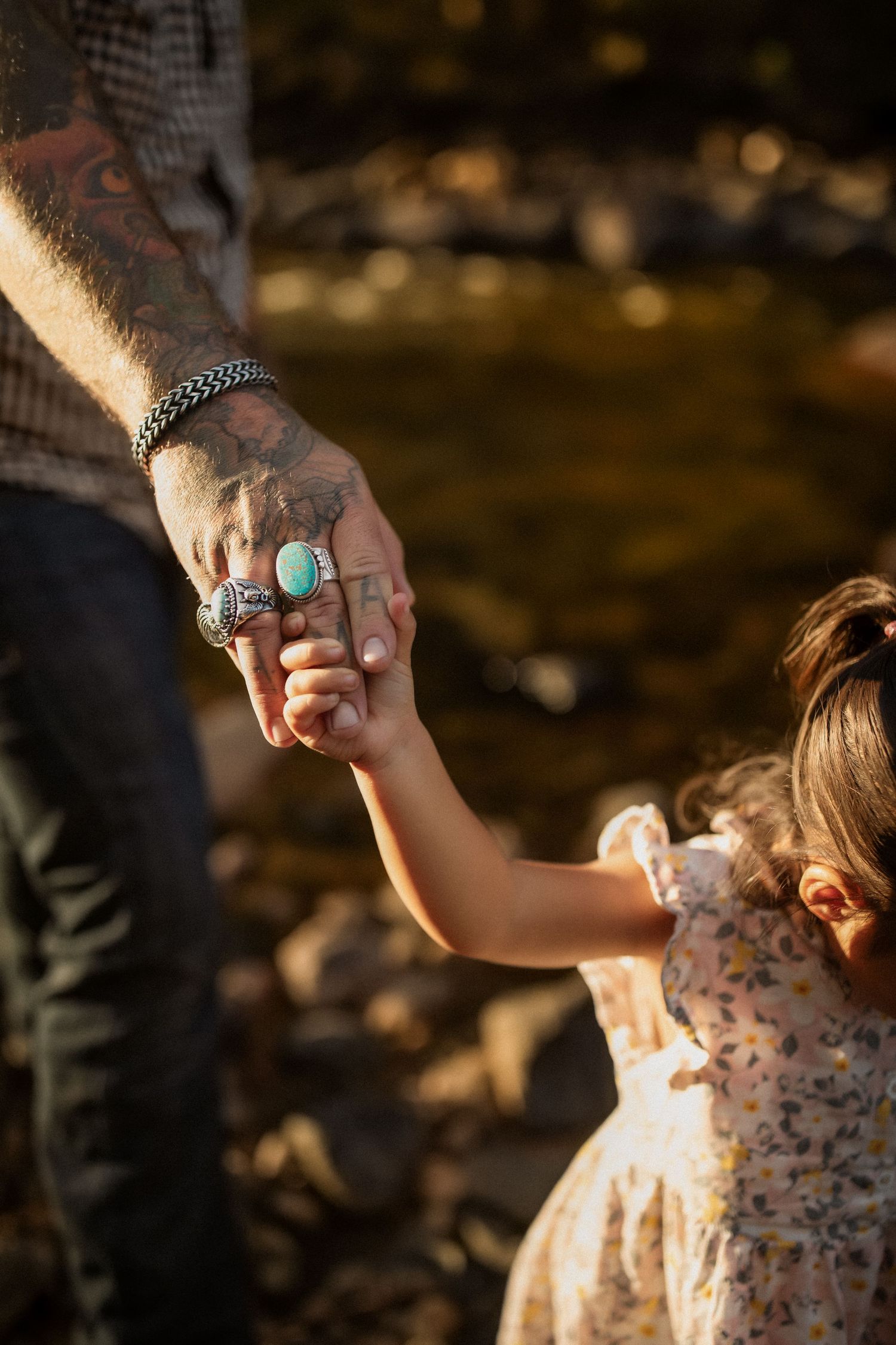 Adult wearing checkered shirt holds child's hand while walking through a sunlit outdoor setting.