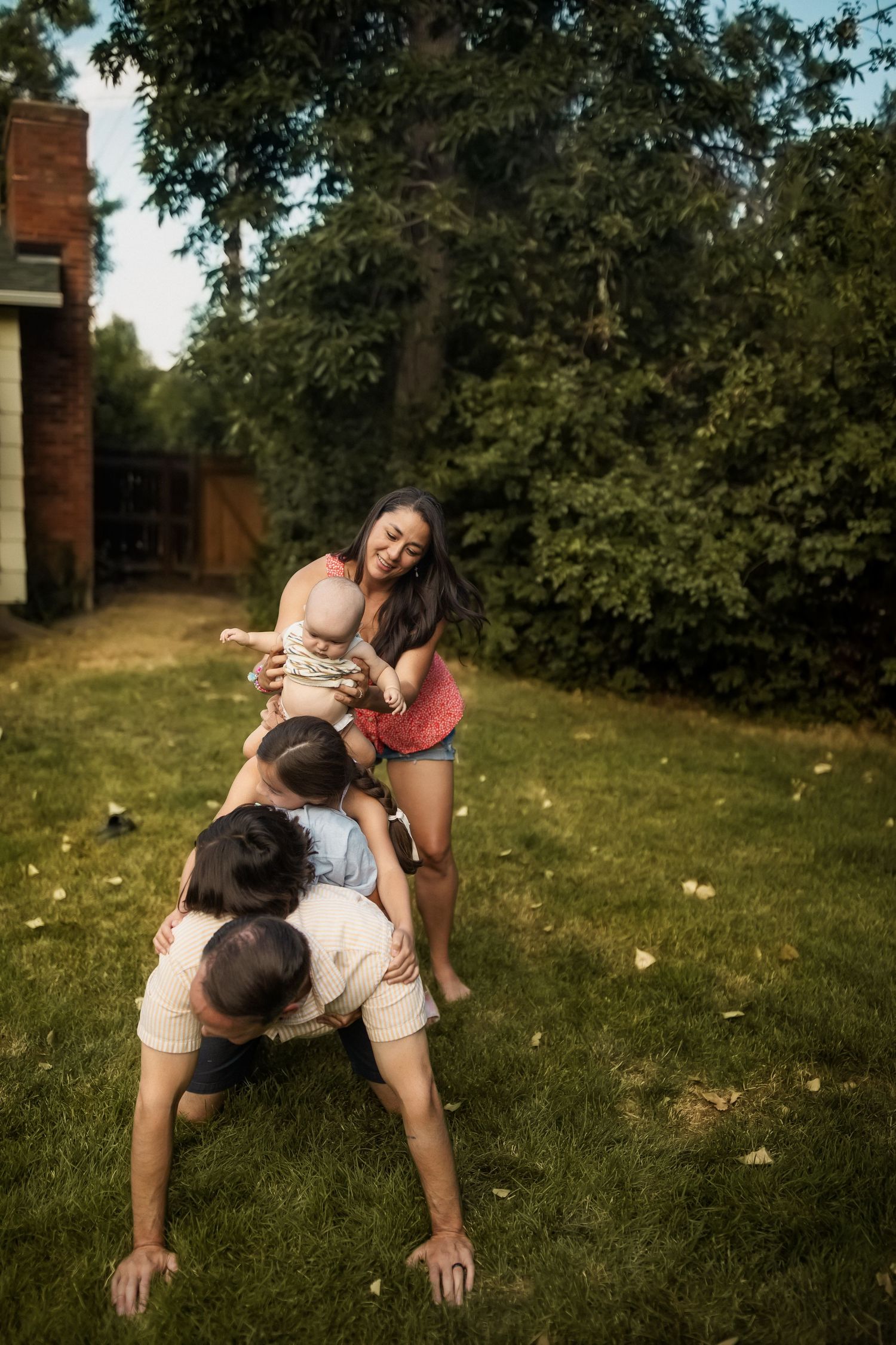 Family playing outdoors in backyard, father on all fours while mother holds baby above him on green grass.
