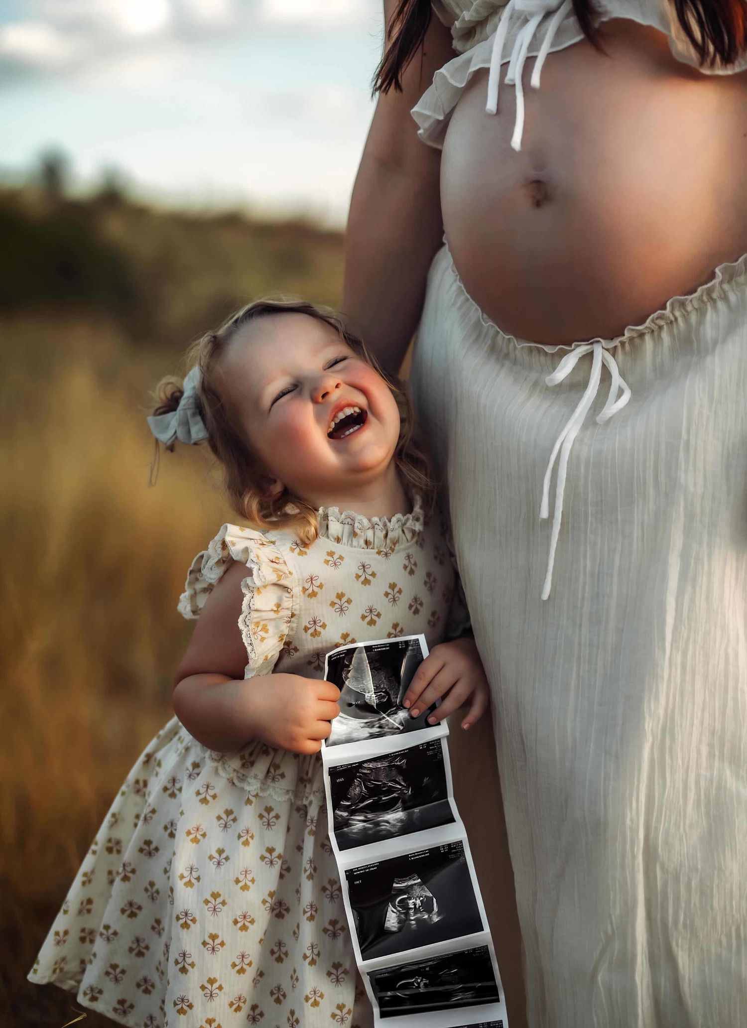A laughing toddler girl holds ultrasound photos beside her pregnant mother in a sunny field, wearing a floral dress.