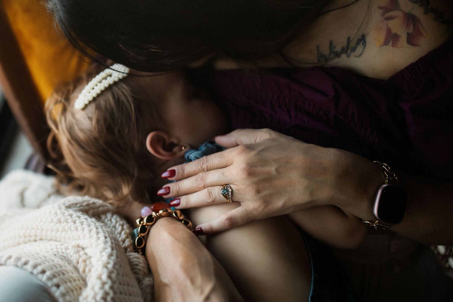 A tattooed mother tenderly breastfeeds her baby, wearing a ring and bracelet, with red manicured nails.