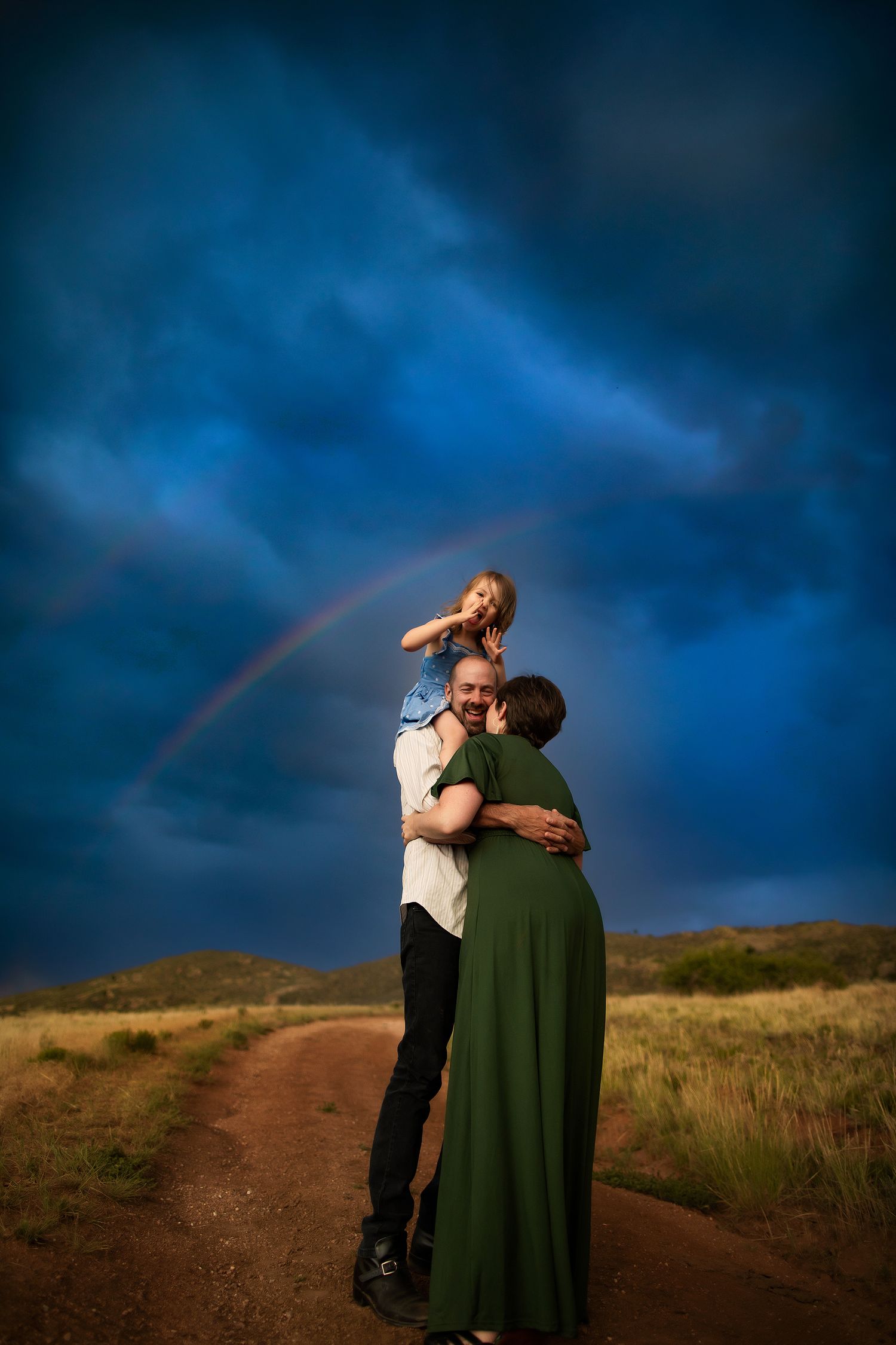 Couple embracing on dirt path with dramatic stormy sky and rainbow in background.