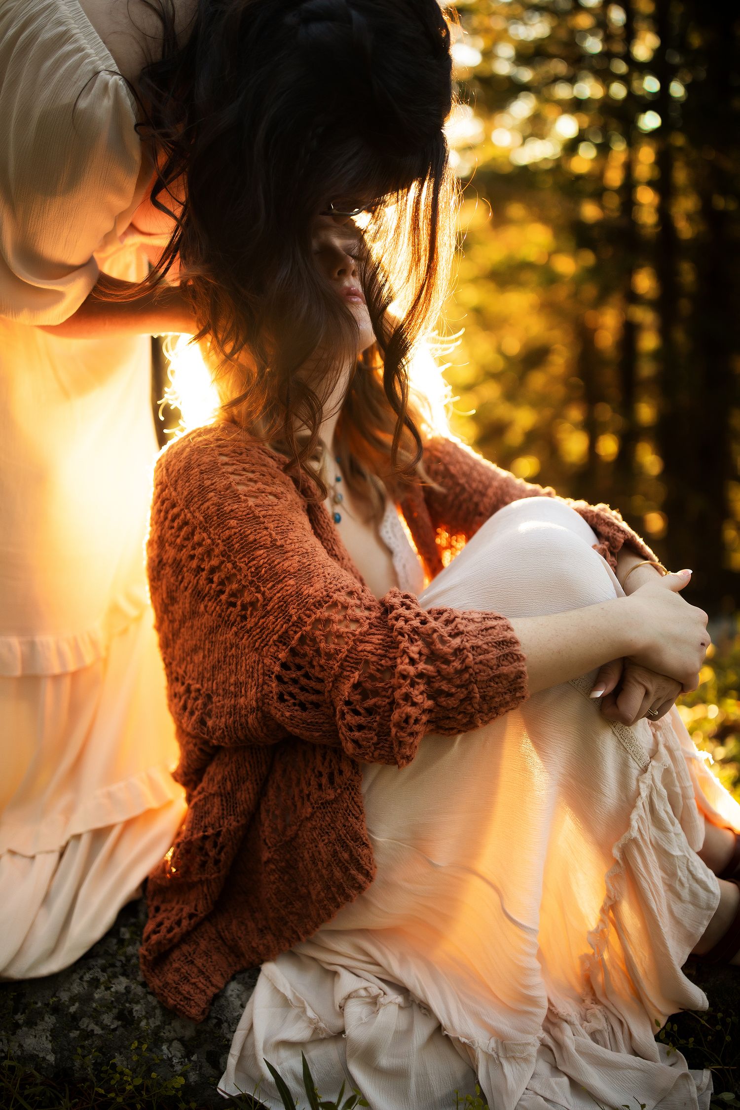 Two women in bohemian attire sit in a sunlit forest, one wearing a rust knit cardigan over a white dress.