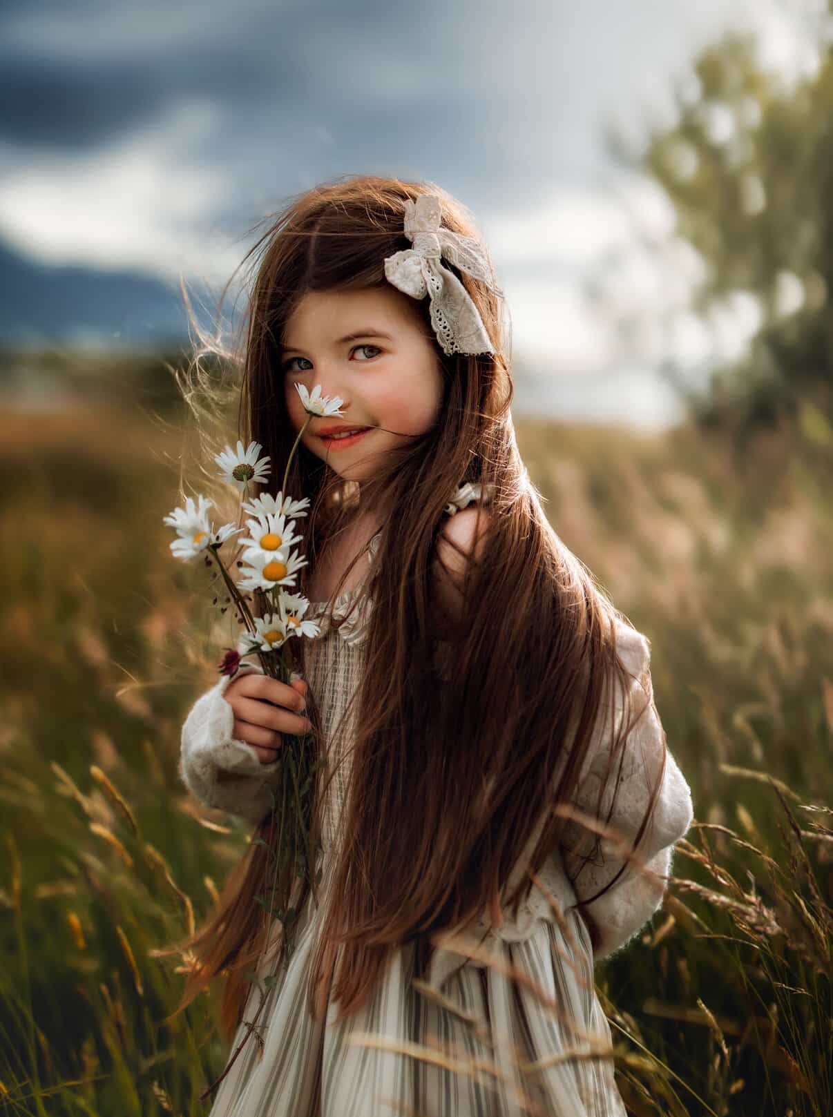 A young girl with long brown hair holds white daisies in a field, wearing a vintage dress with a bow hair clip.