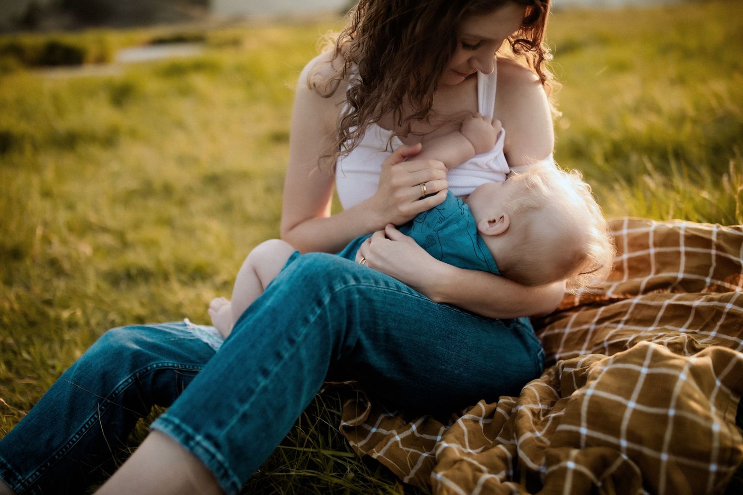 Mother breastfeeding baby outdoors on plaid blanket in green field during golden hour sunset.