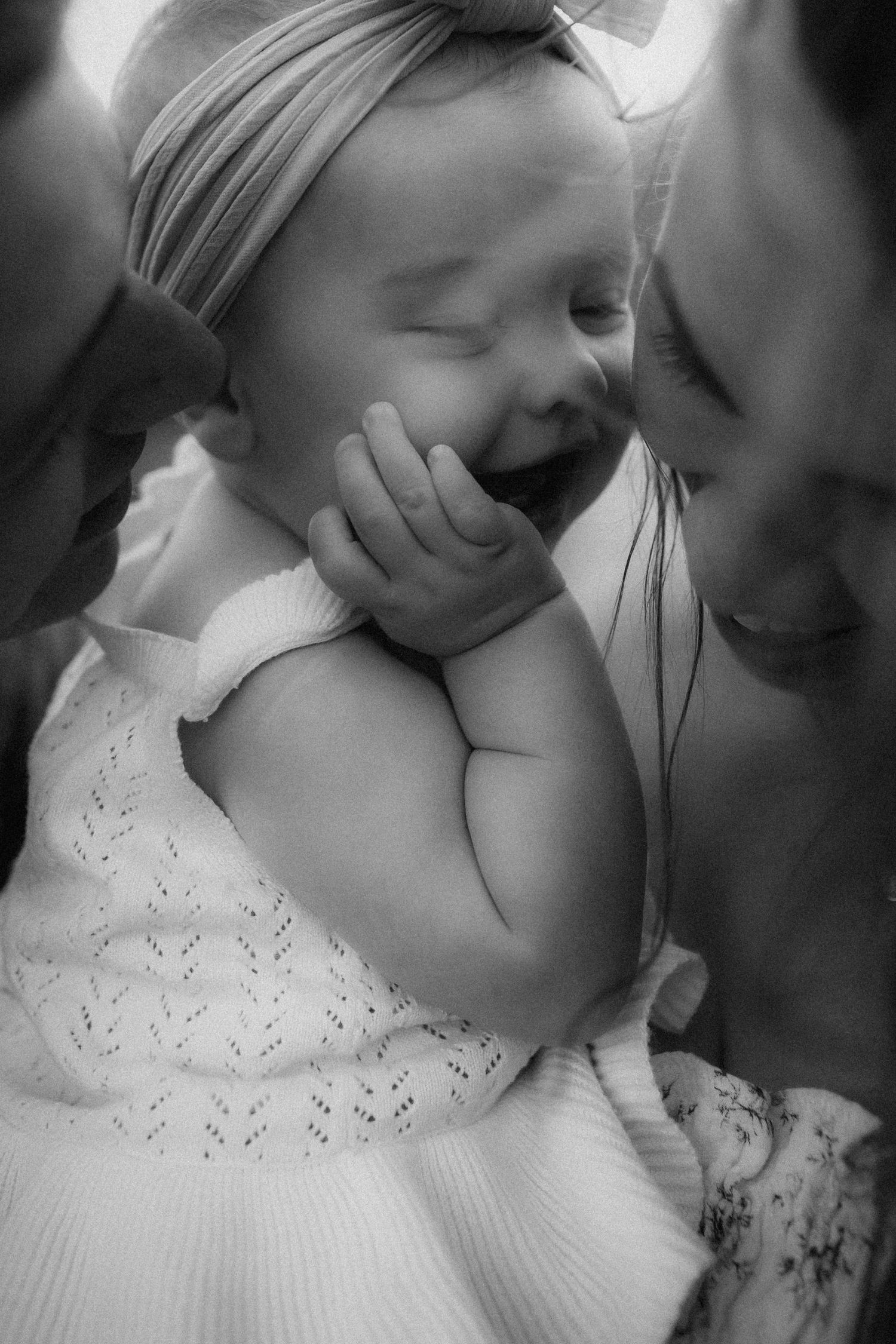 Black and white photo of a laughing baby in white dress with bow headband, held by parents on each side.
