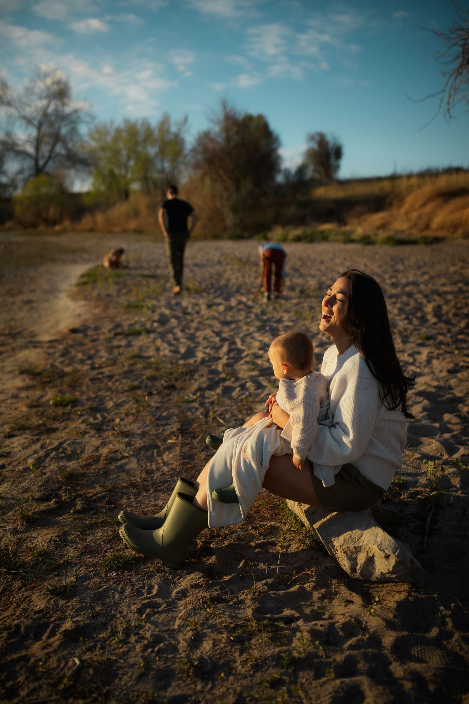 A mother sits on sandy ground holding her baby in a white wrap, wearing green boots, with family members in the background.