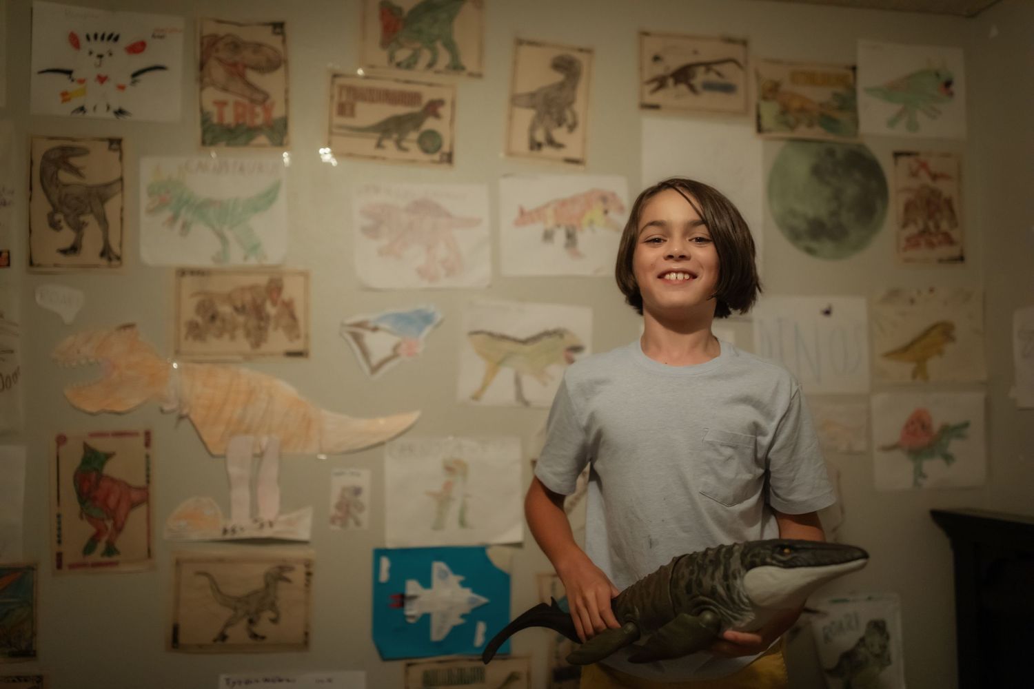 A student stands in front of a wall covered in dinosaur drawings and illustrations in a warmly lit room.