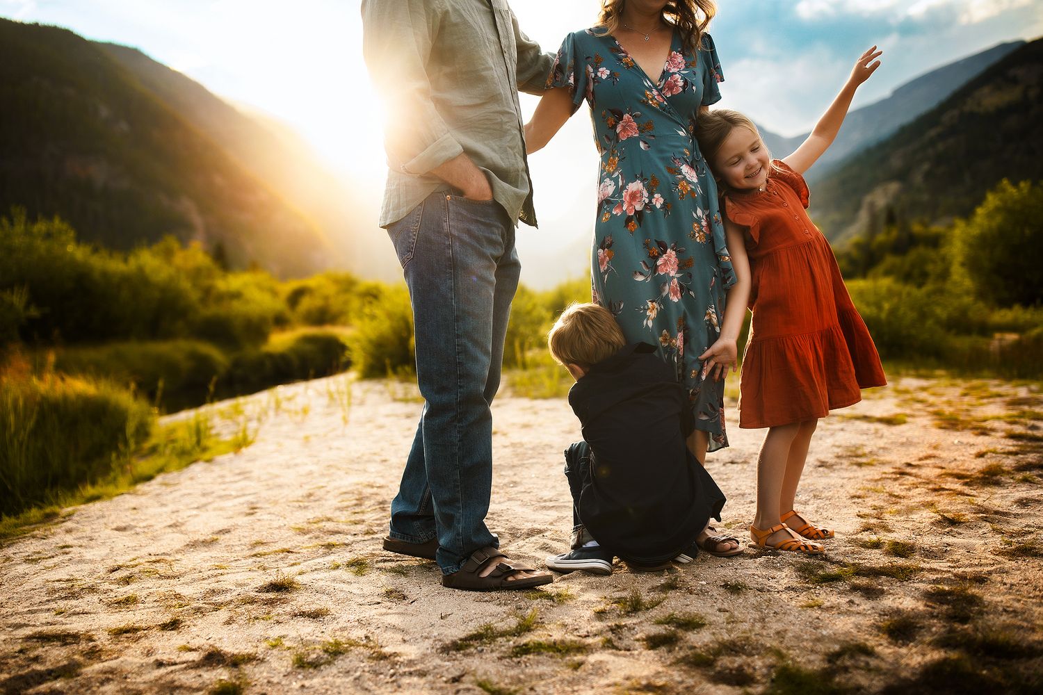 People's feet and legs stand on sandy ground during golden sunset in a mountainous landscape.