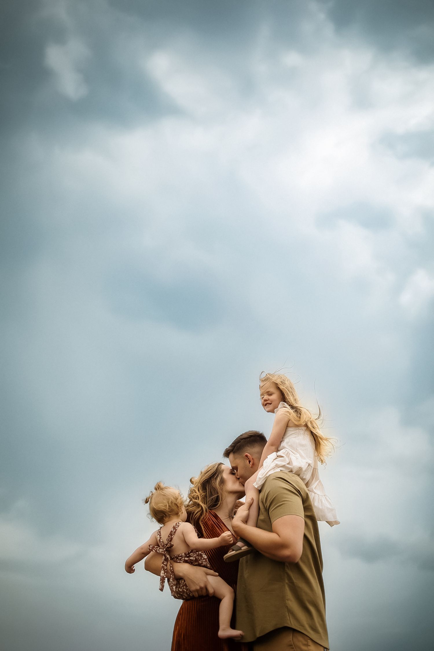 Family embraces against dramatic cloudy sky in heartwarming outdoor portrait series.