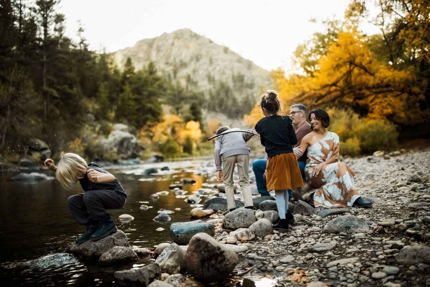 Family gathering on rocky riverbank surrounded by autumn foliage and mountain scenery.