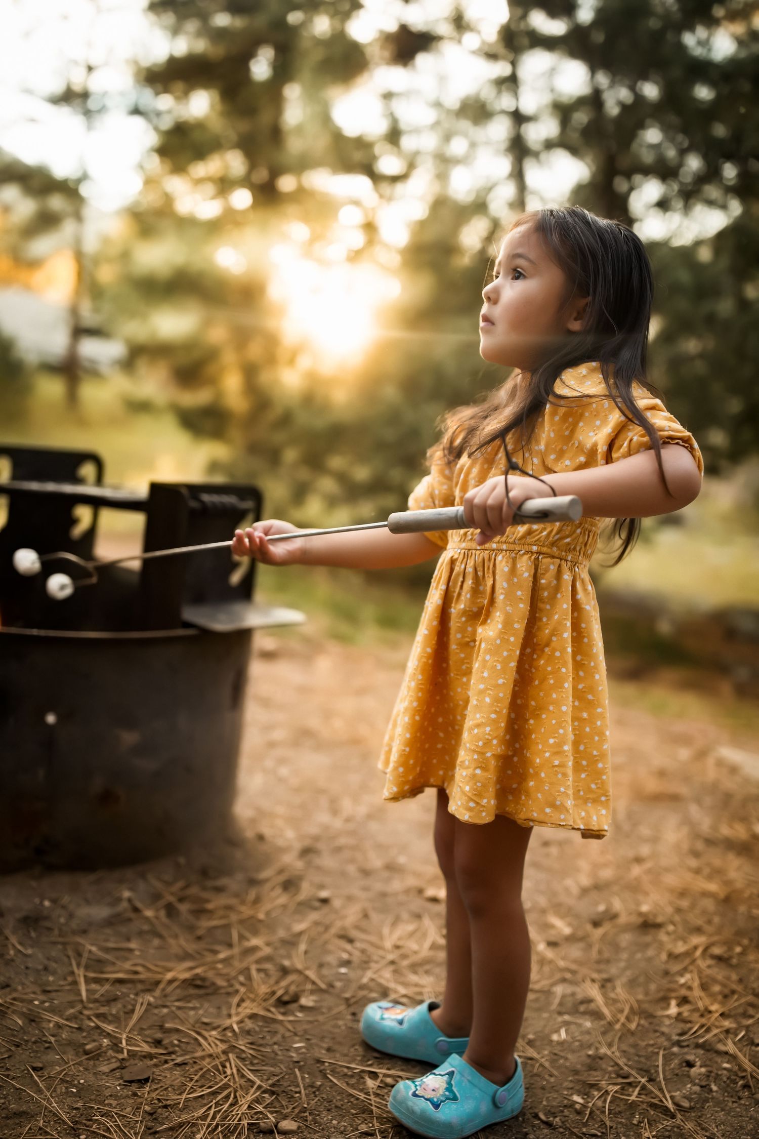 Young person in yellow dress roasting marshmallows outdoors at golden hour.