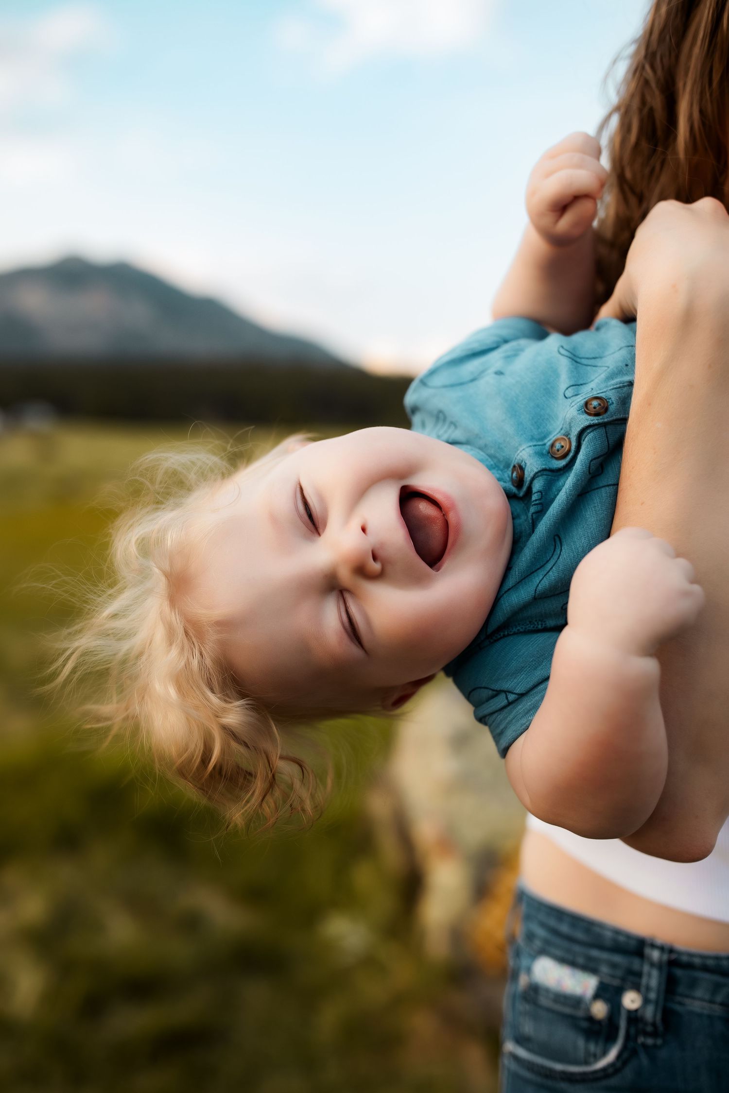 A joyful toddler laughs while being held upside down by a mother outdoors with mountains in the background.