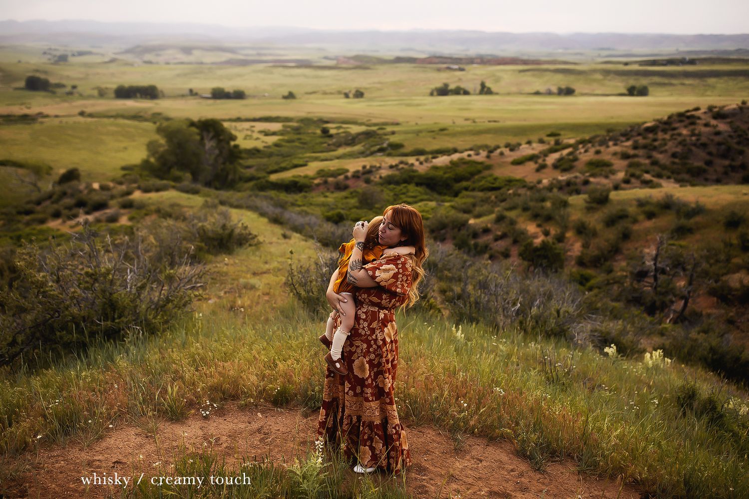 Two figures share an intimate moment on a dirt path overlooking expansive grasslands at sunset.