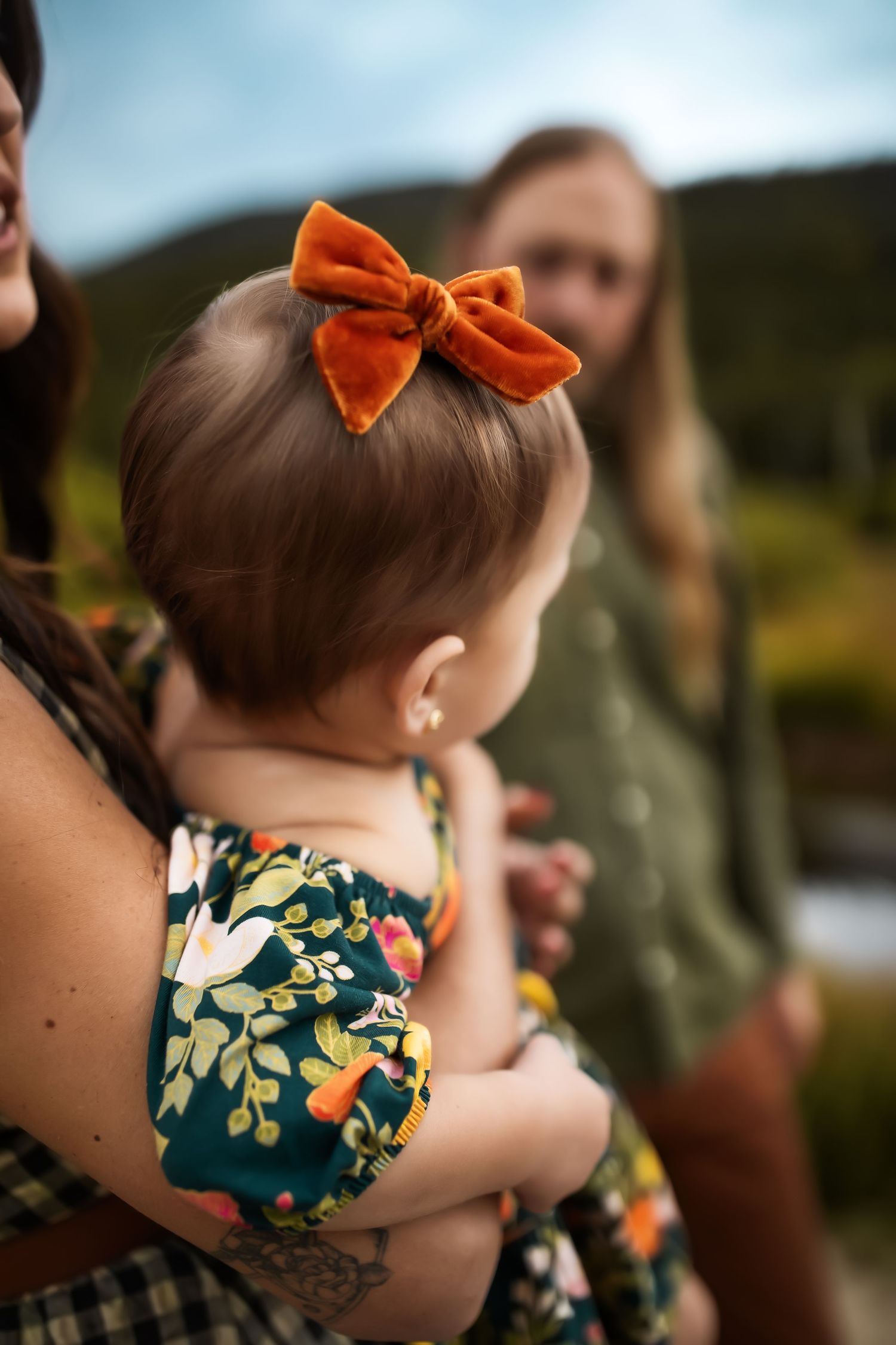 A child with a bright orange bow in their hair wears a floral patterned dress outdoors.